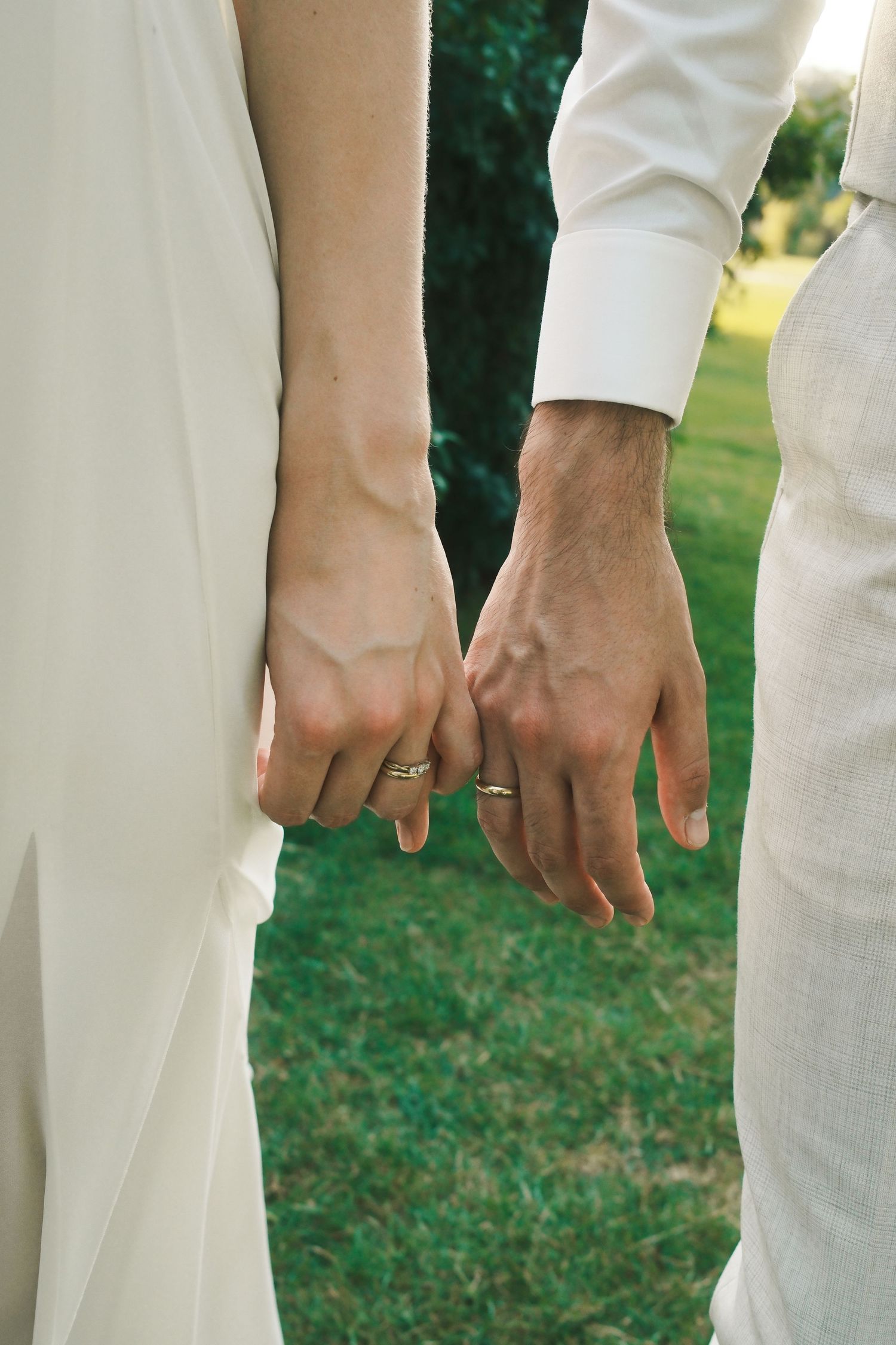 Newlywed couple holding hands outdoors, showing gold wedding rings, wearing white wedding attire on green grass.