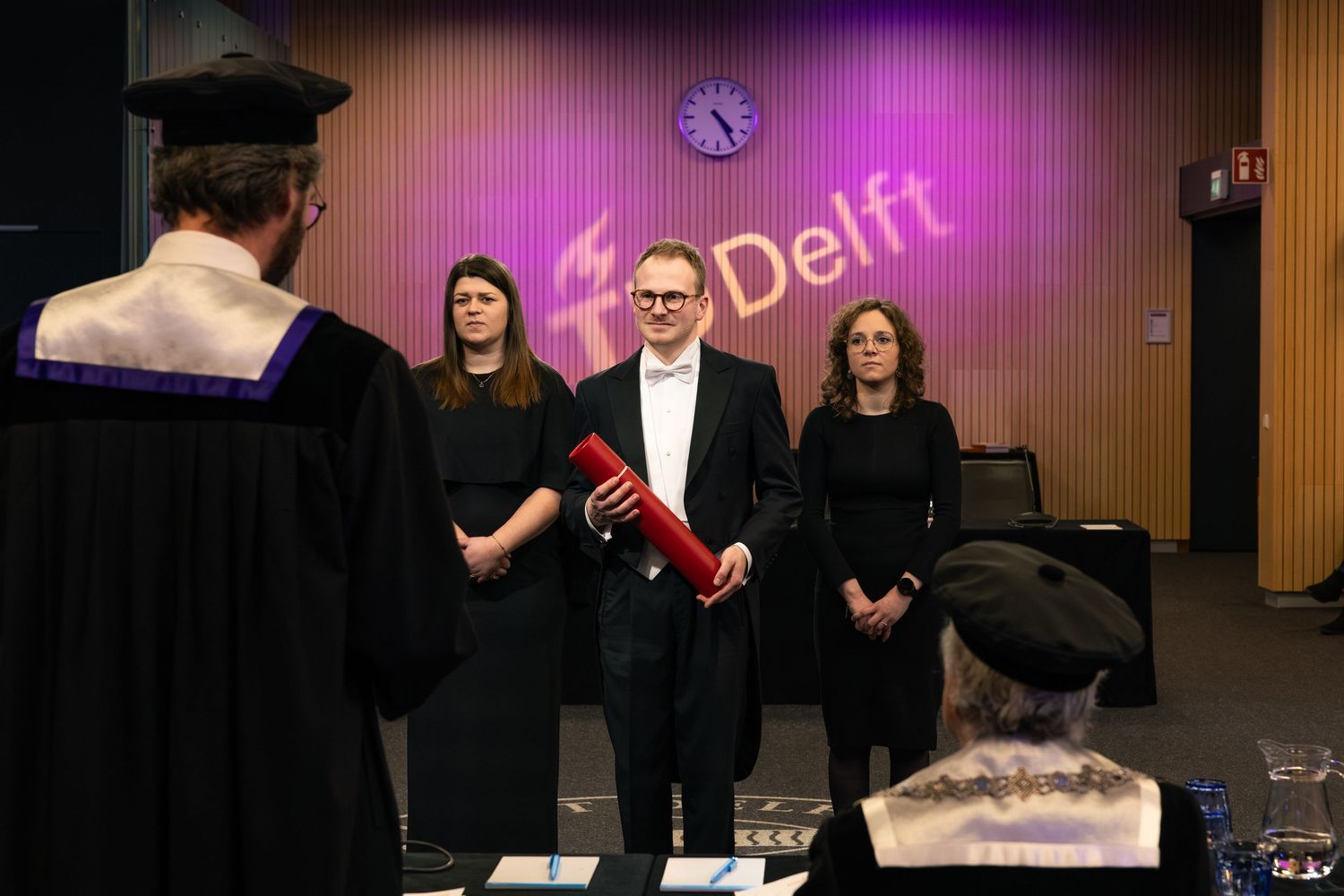 PhD ceremony at TU Delft with graduate holding diploma, flanked by two women in academic hall.