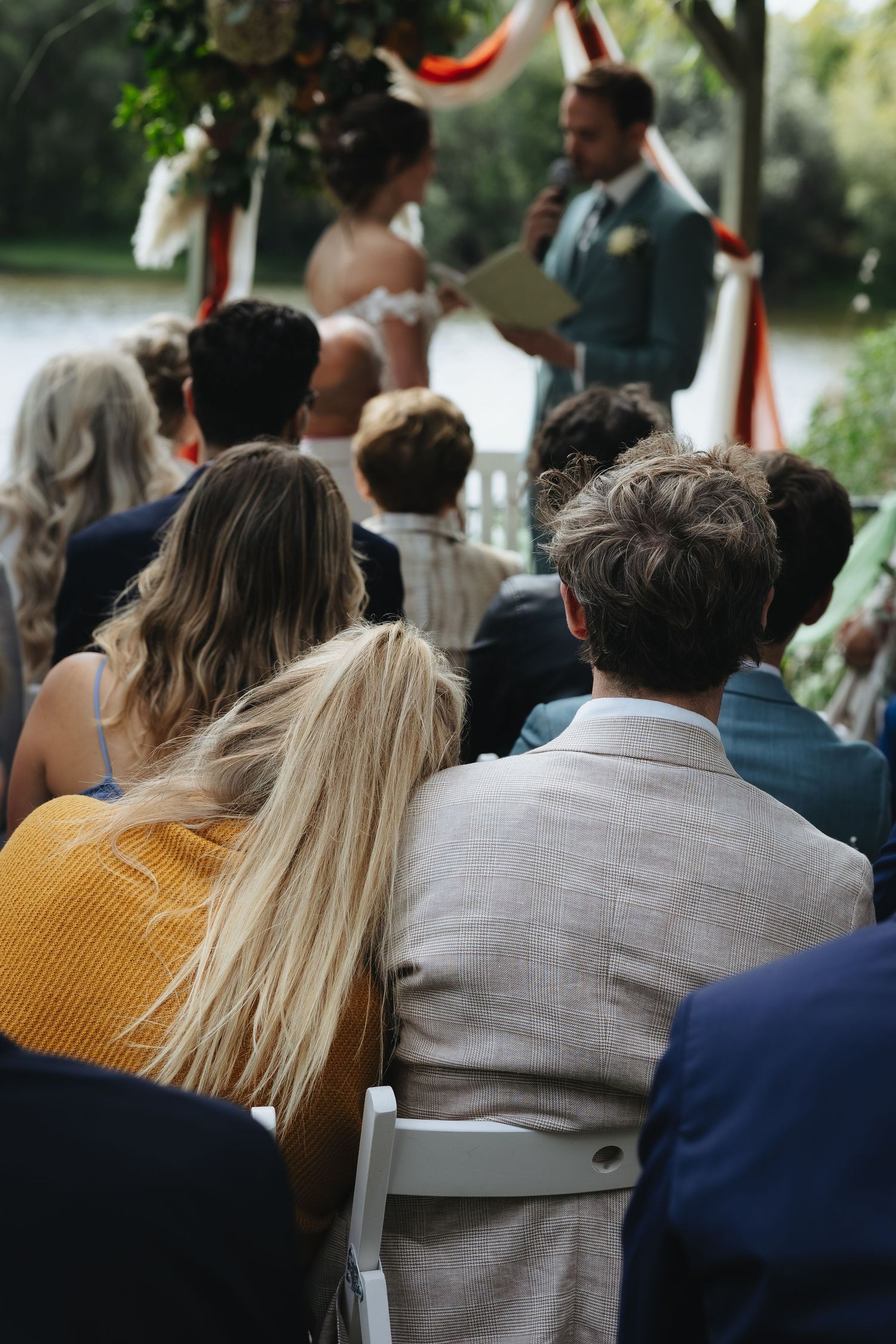 Guests seated outdoors at an elegant wedding ceremony, watching couple exchange vows near a floral arch by a lake.
