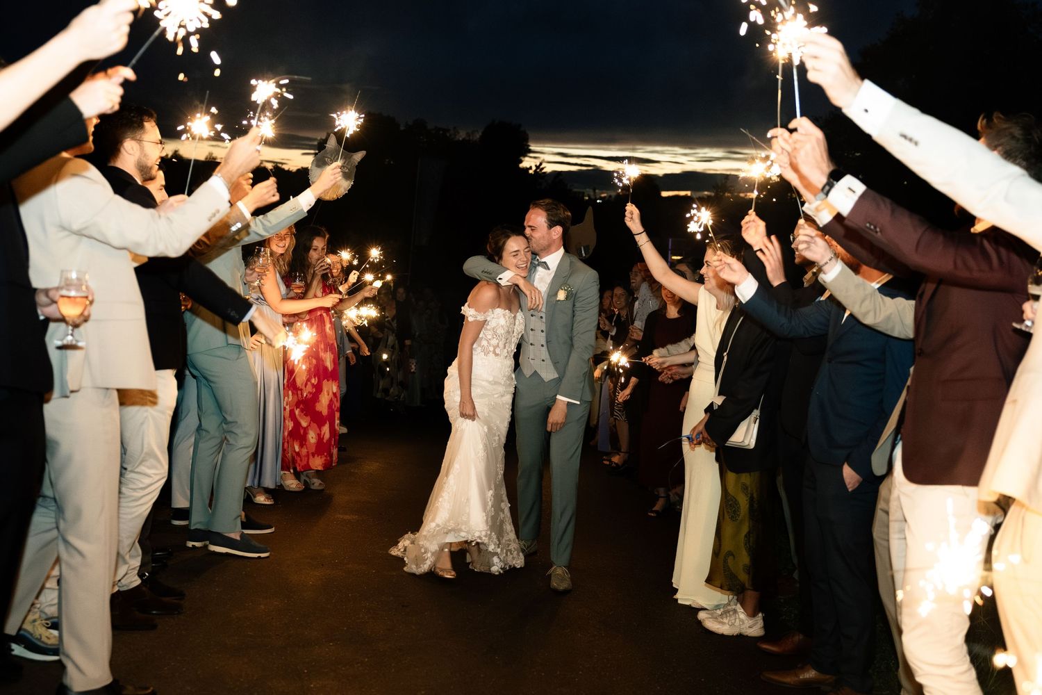 Bride and groom walk through sparkler tunnel surrounded by cheering wedding guests at night.