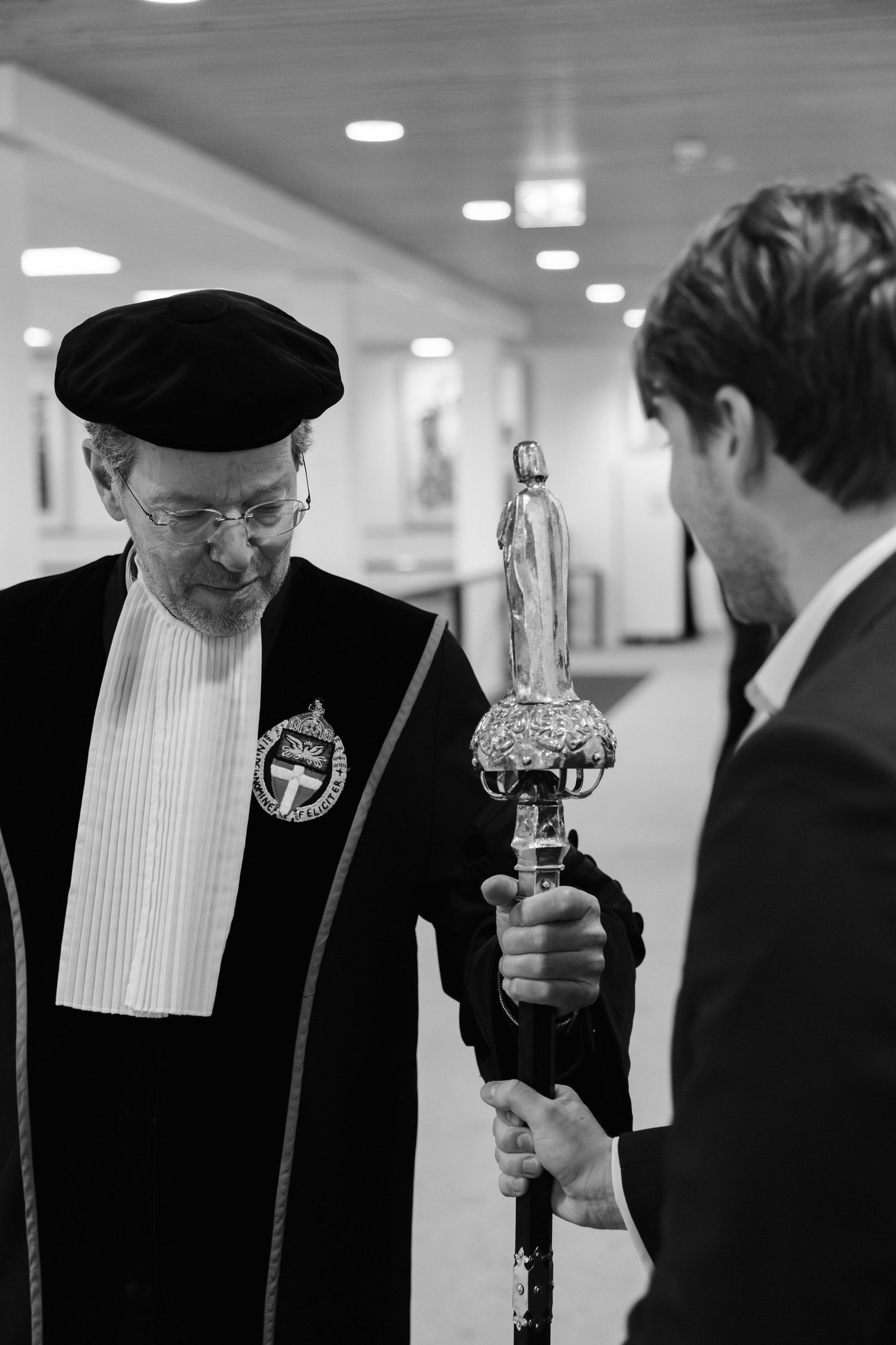 Black and white photo of an academic in ceremonial robes and cap examining an ornate ceremonial mace held by a suited man.