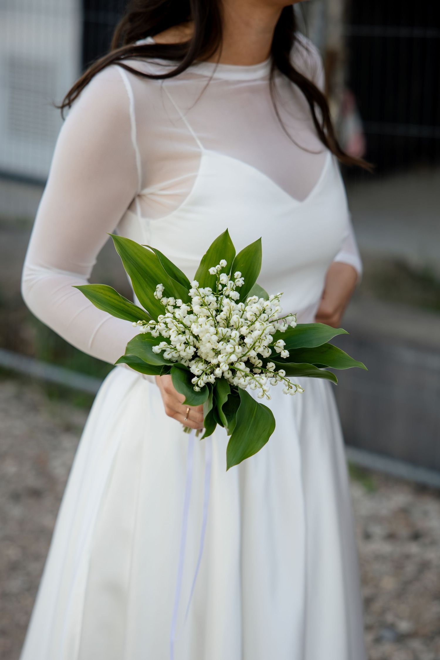 Bride in white mesh dress holding elegant lily of the valley bouquet with green leaves at wedding ceremony.