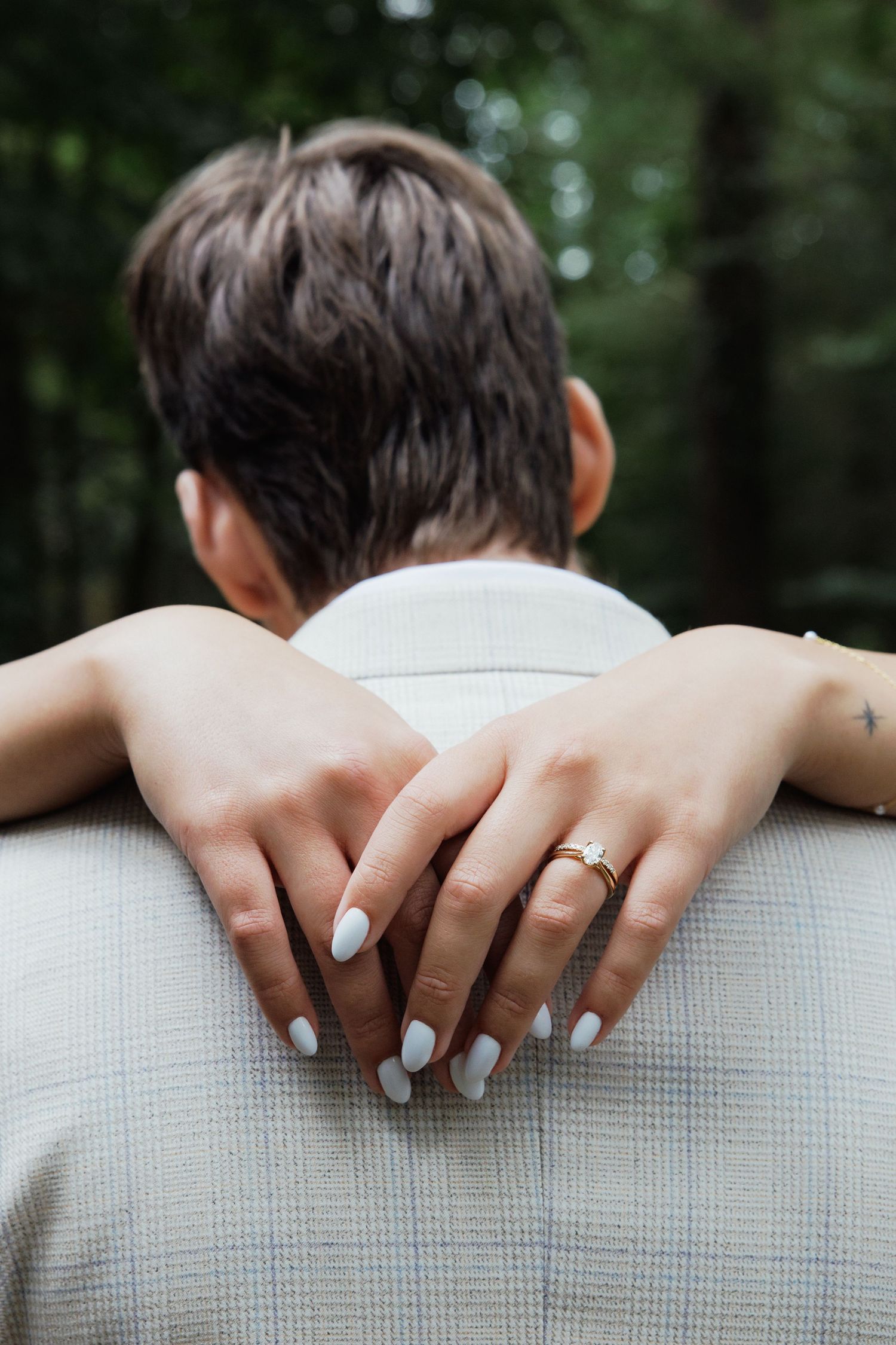 A person with short hair being embraced from behind, showing hands with white nails and a gold engagement ring.