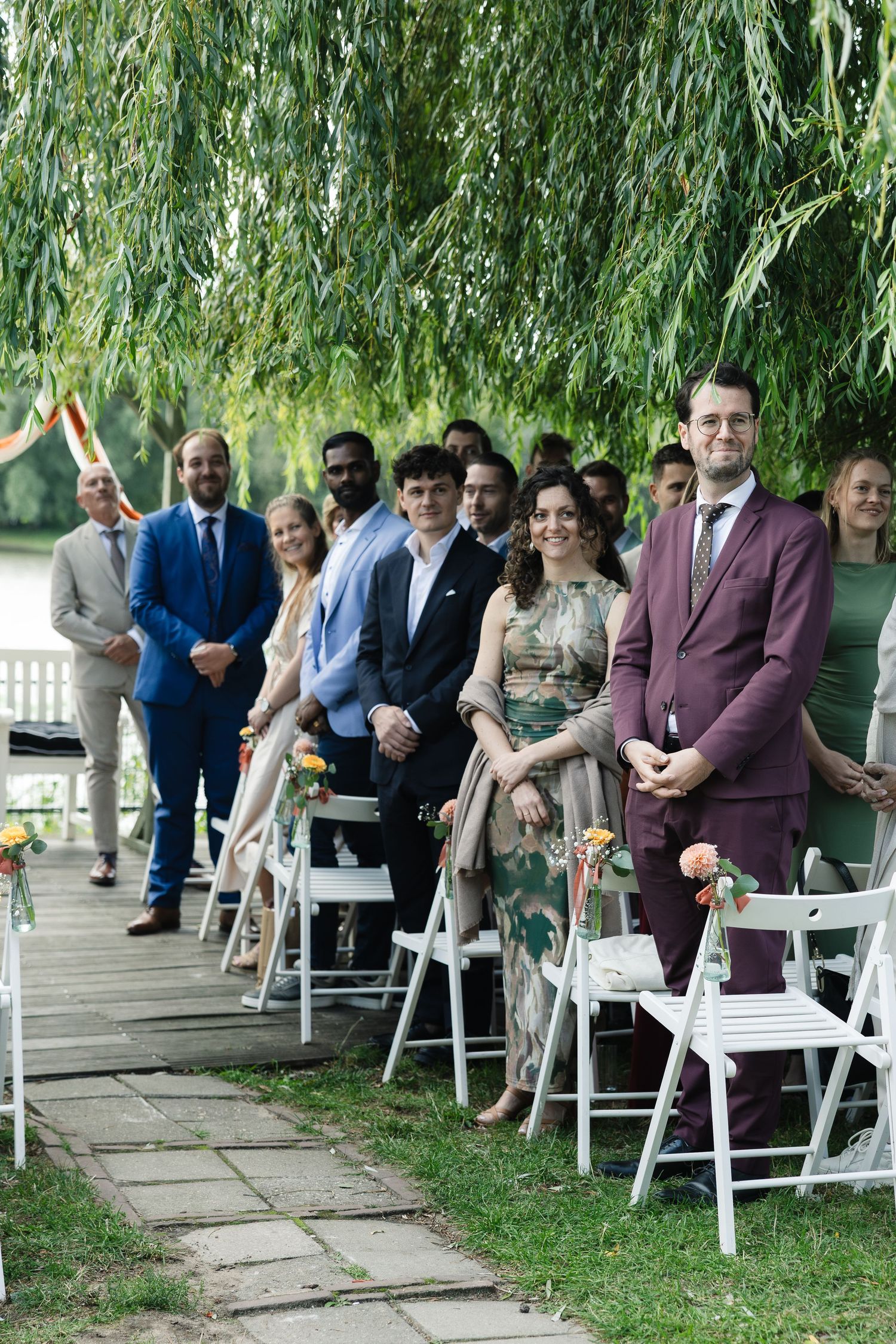 Guests standing and seated in white chairs at an outdoor wedding ceremony under a weeping willow tree.