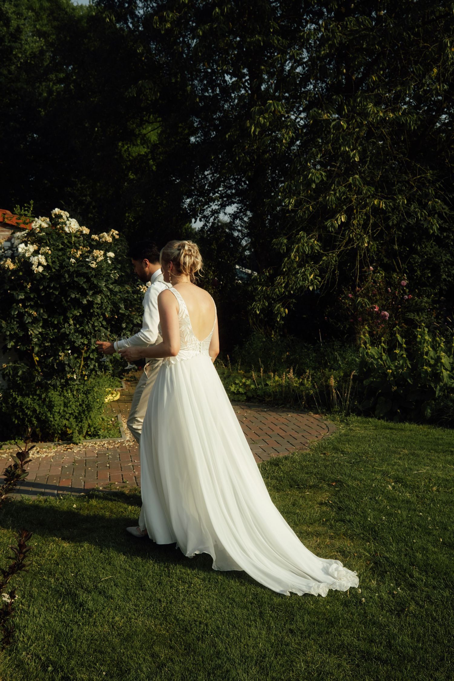 Bride in elegant backless white gown walks with groom through lush garden path at golden hour wedding.