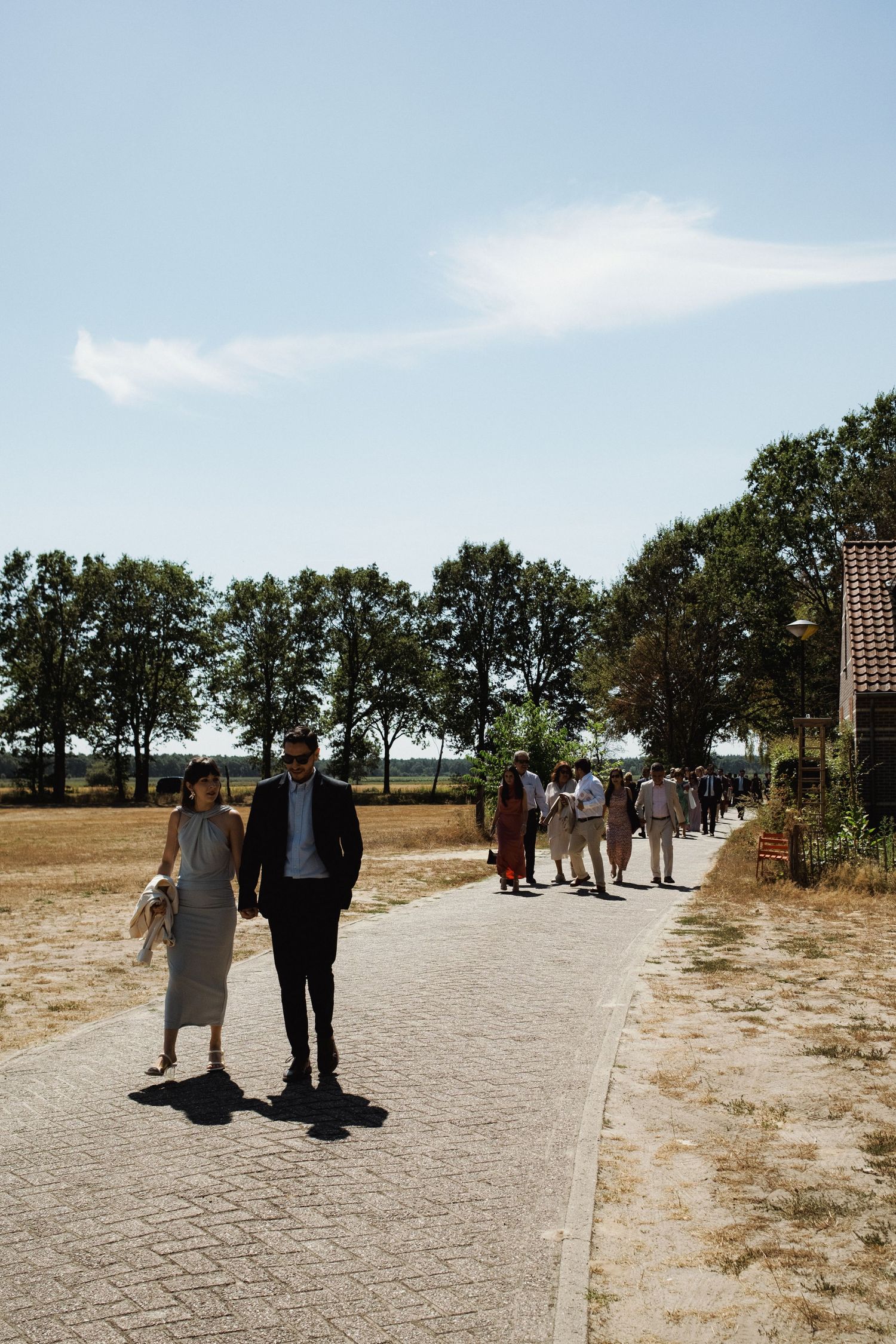 Couple walking on a cobblestone path with guests and trees in the background on a sunny day.