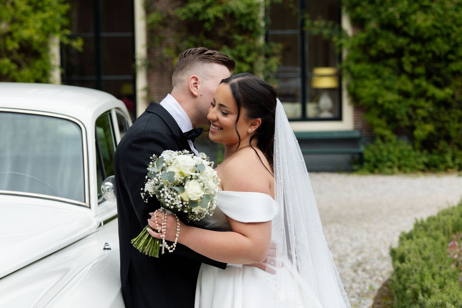 Bride and groom embracing near a vintage white car, bride holding a white bouquet and wearing a veil.