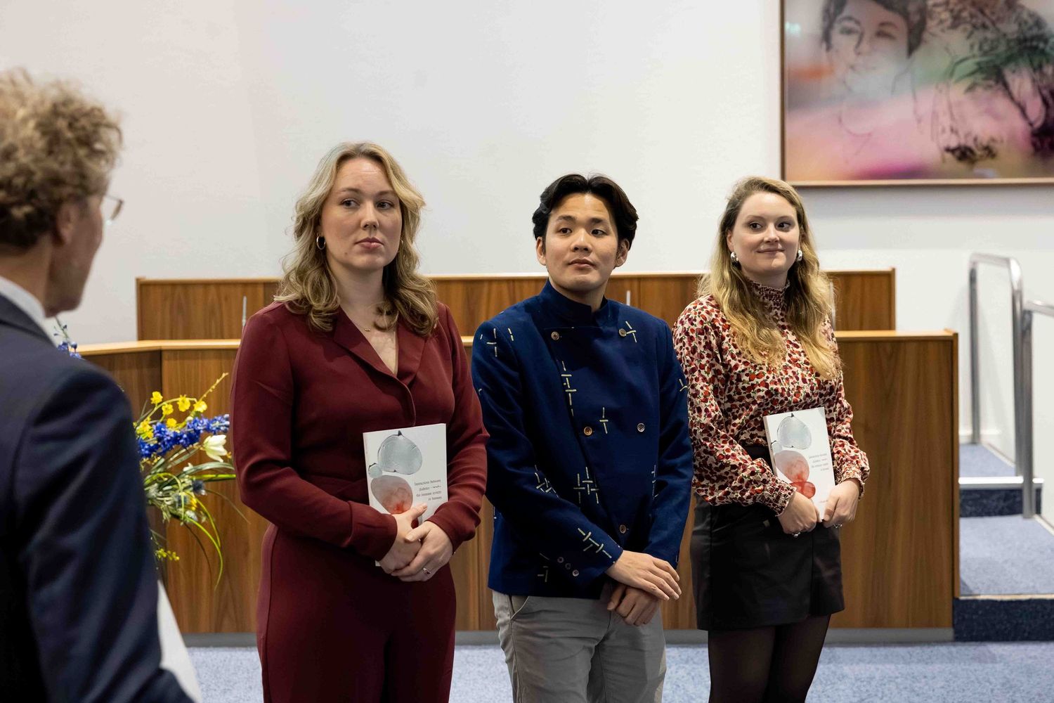 Three people holding books stand in a formal indoor setting during an award or recognition ceremony.