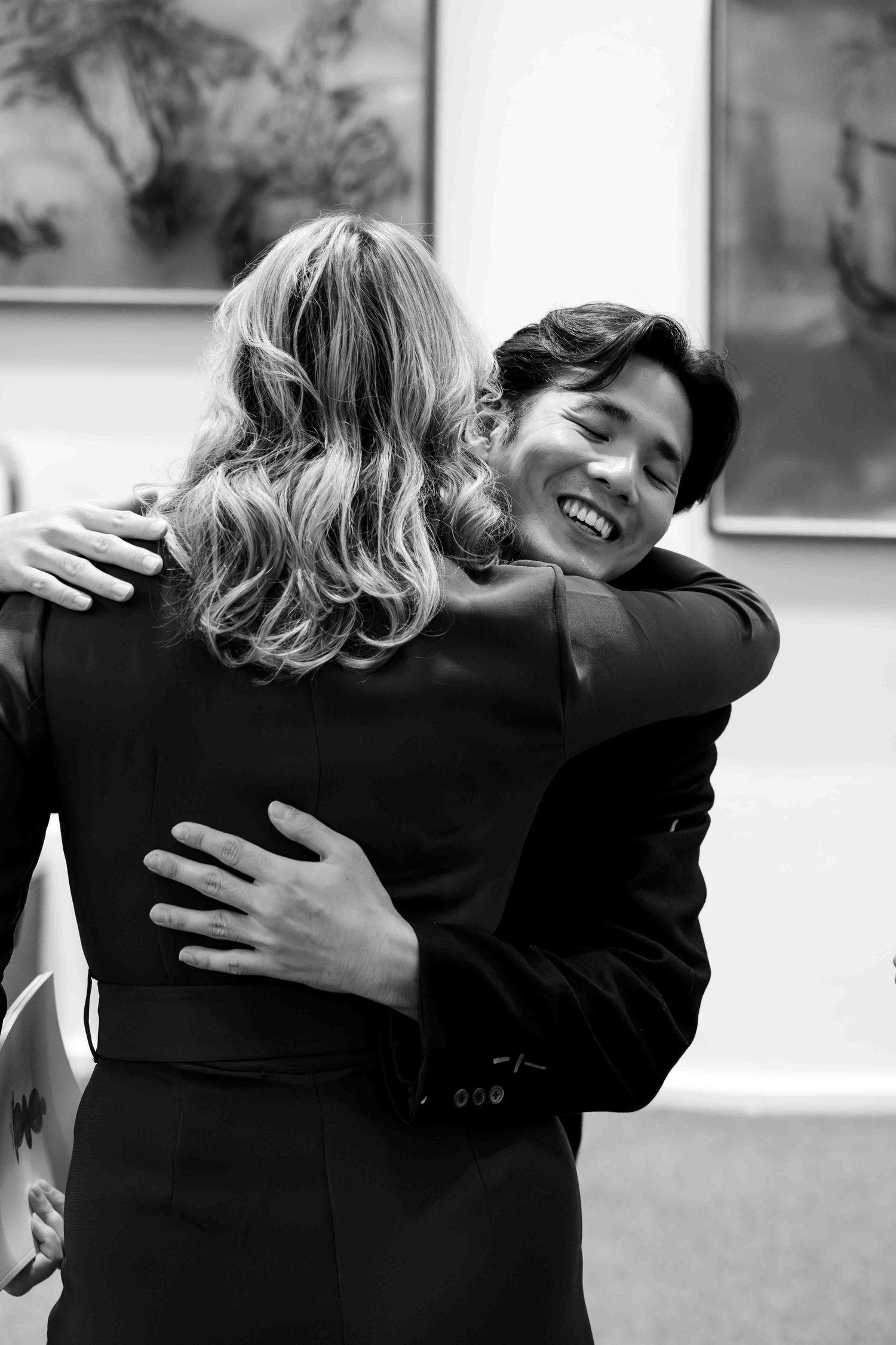 Two people in formal attire share a warm embrace, smiling, in a black and white photo.