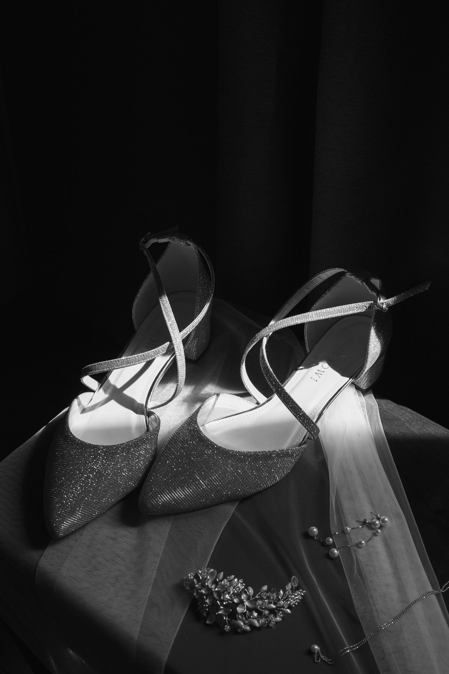 Black and white photo of glittery bridal heels with ankle straps resting on a veil beside floral hair accessories.