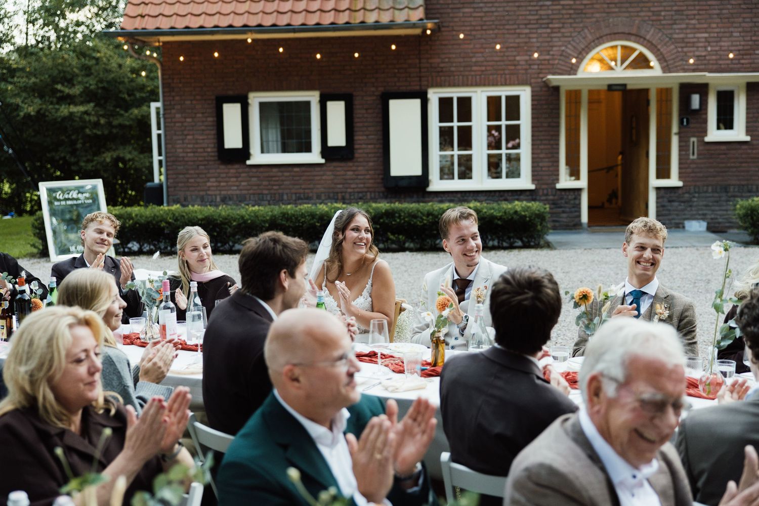 Outdoor wedding reception with guests seated at decorated tables near a brick house with string lights.