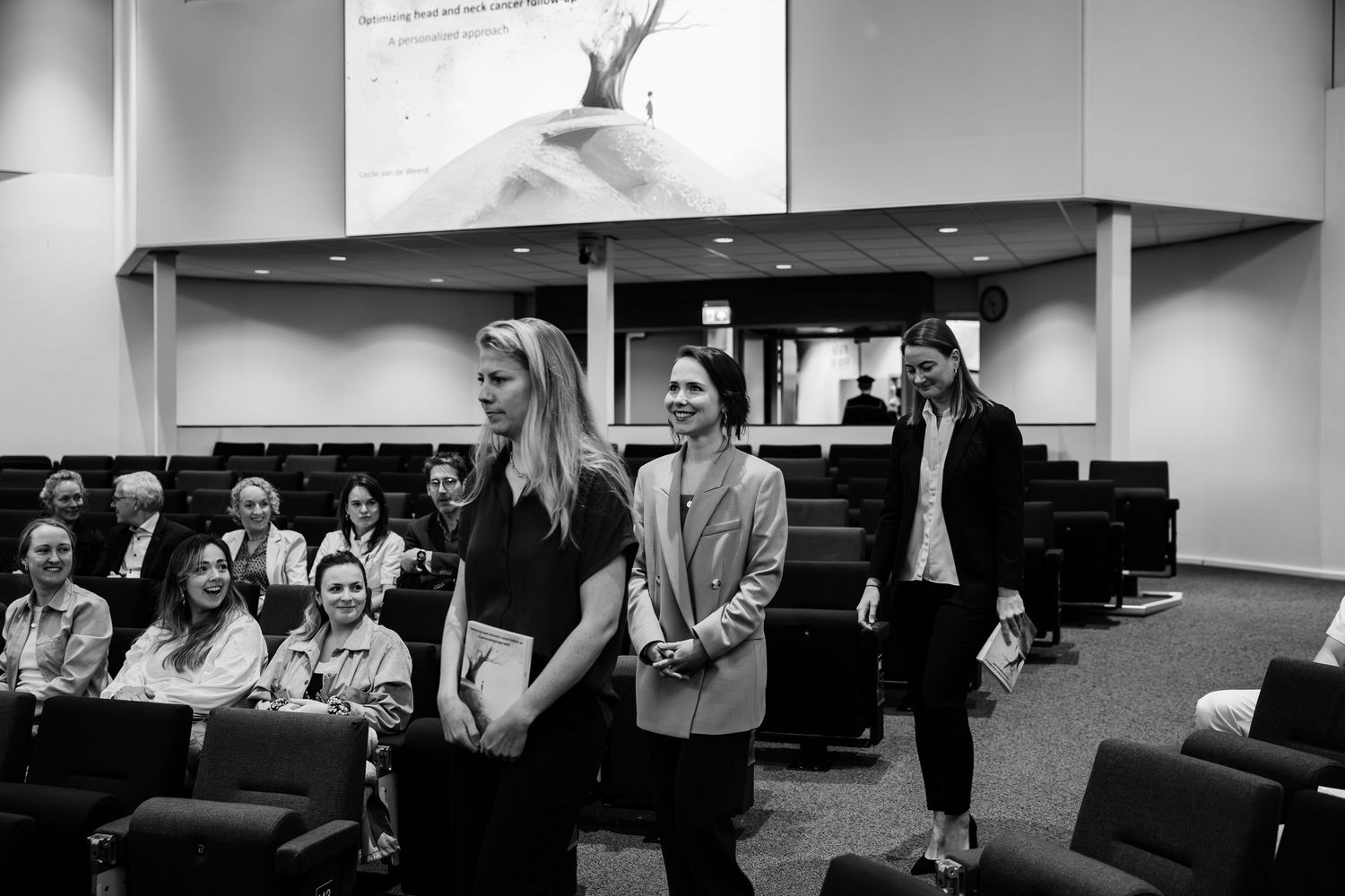 Three women stand in a modern conference room during a professional presentation event with audience seated in background.