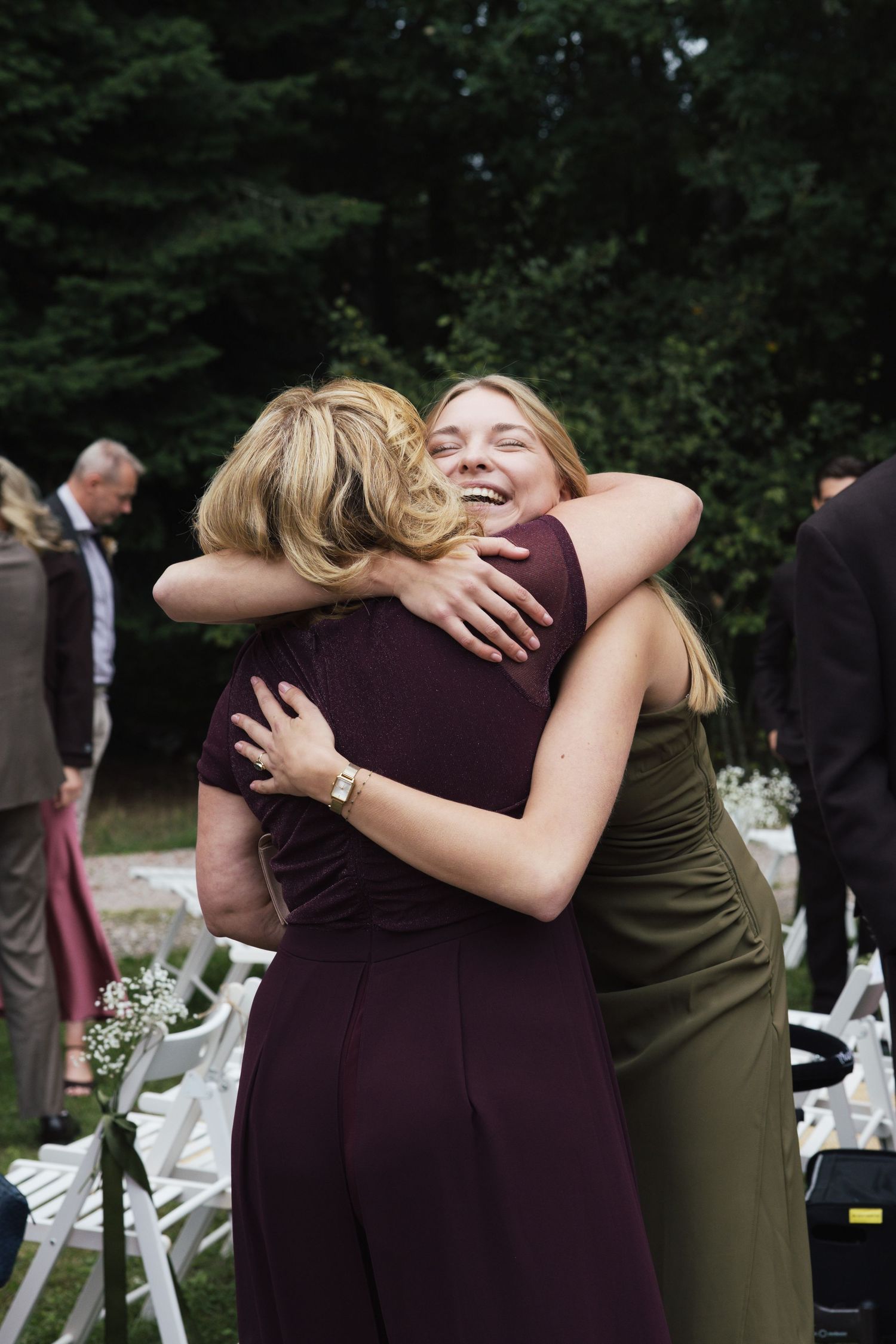 Two women sharing a warm hug at an outdoor wedding ceremony, wearing elegant maroon and olive dresses.