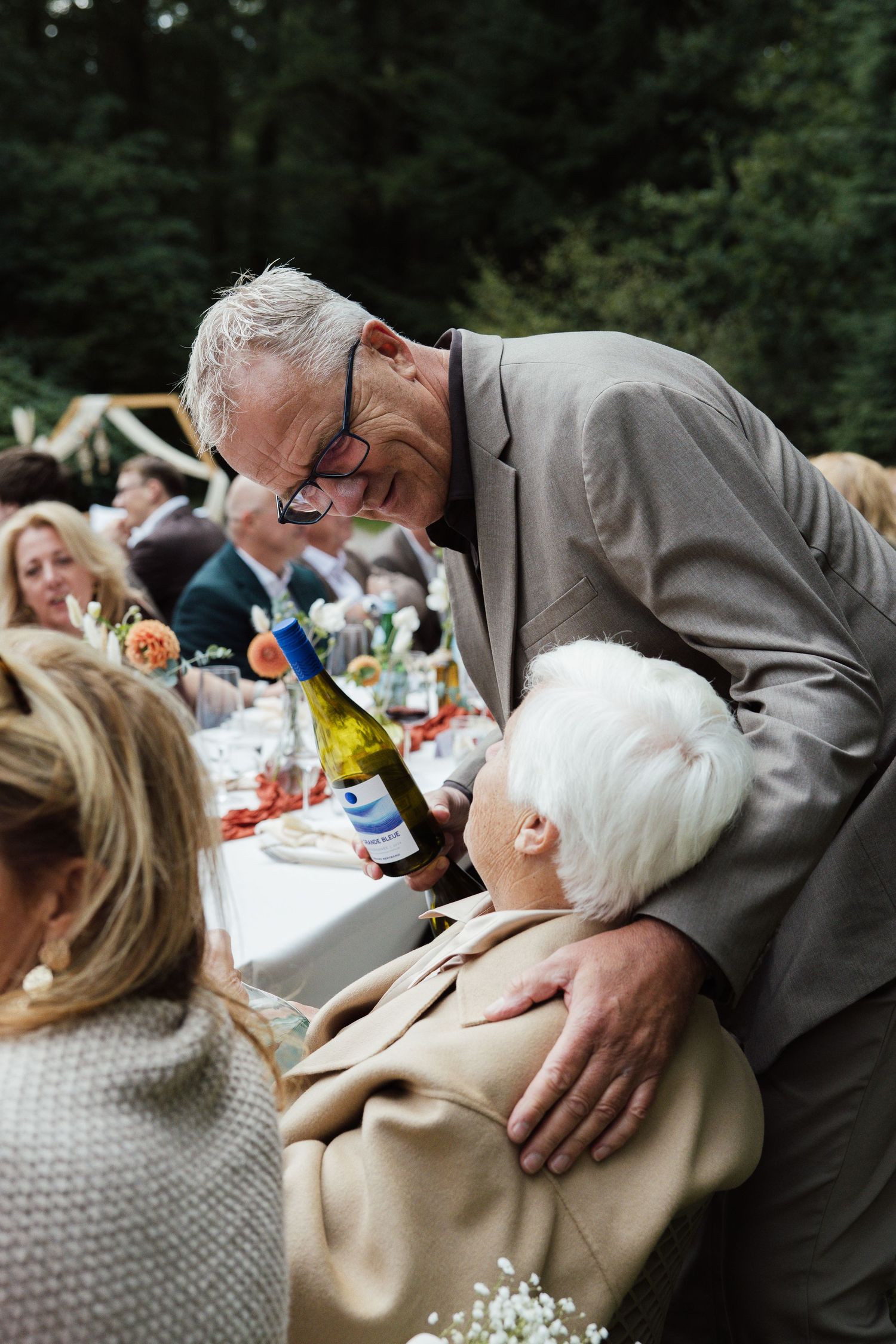 Elderly man in gray suit smiling and holding wine bottle at outdoor wedding reception dinner party.