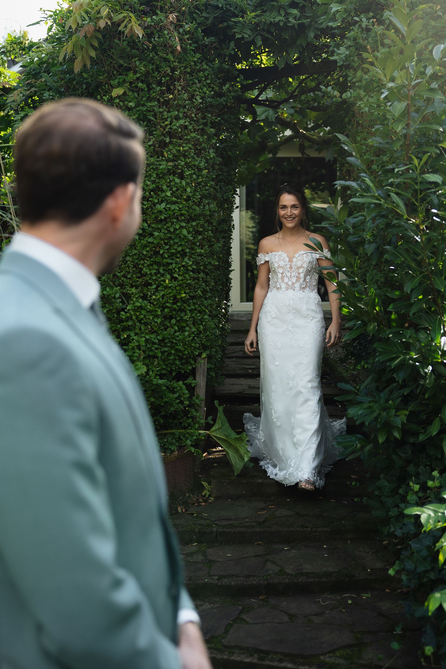 Bride in lace off-shoulder gown walks through garden arch toward groom in sage suit during outdoor wedding.
