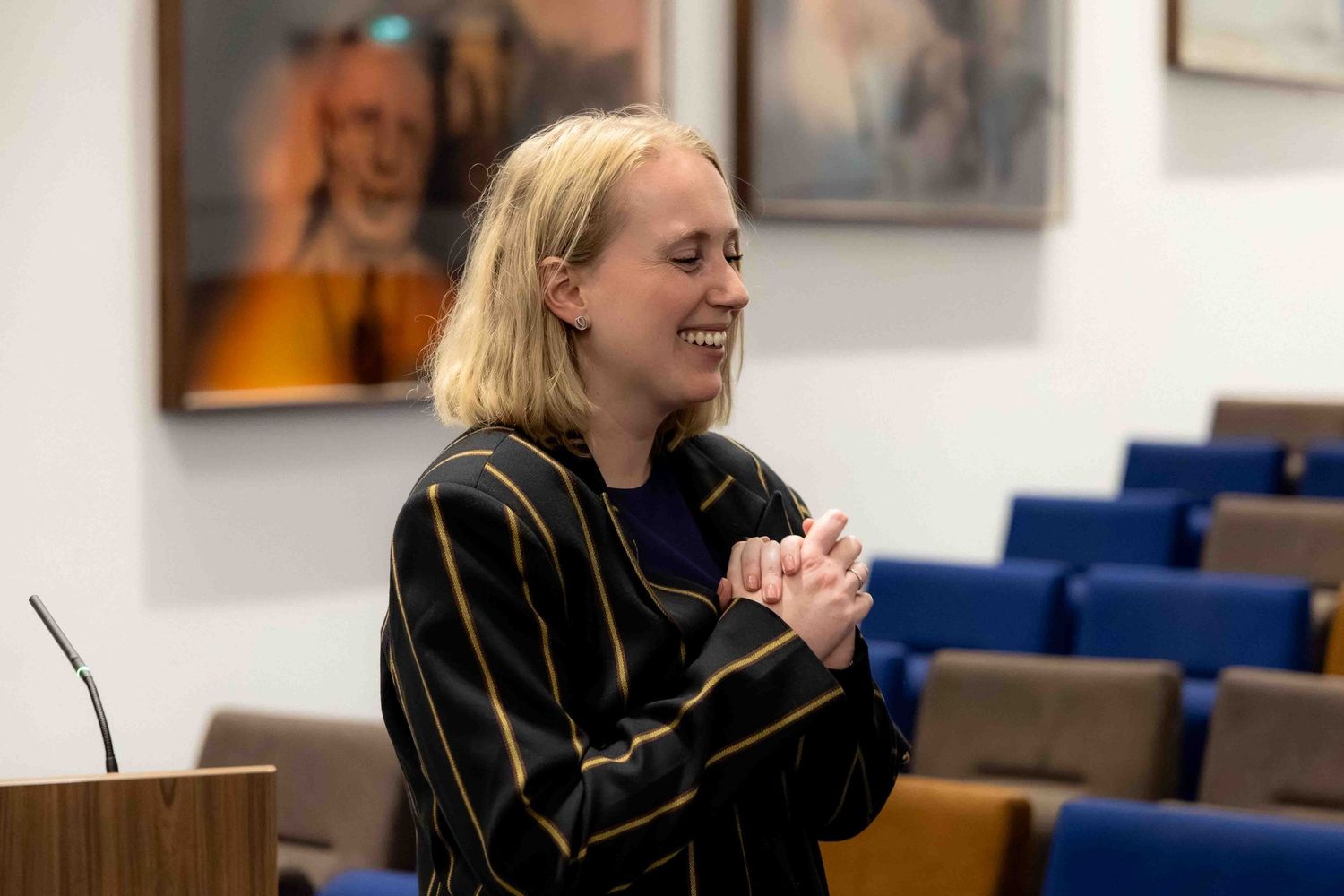 Smiling blonde woman in striped jacket clasping hands in a conference room with blue chairs and portrait painting.
