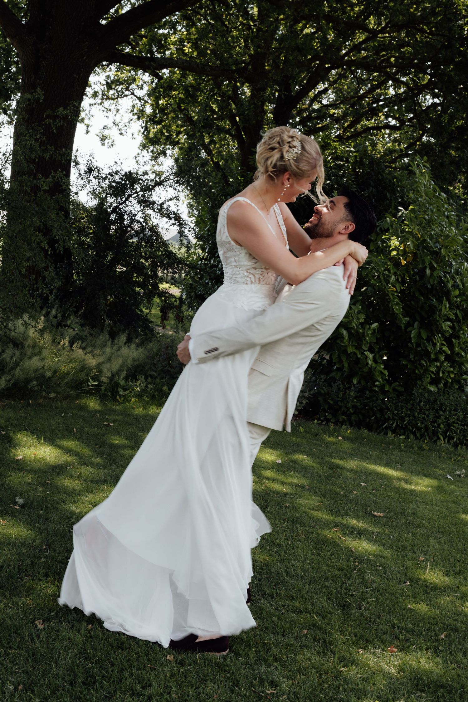 Groom lifts bride in white wedding dress outdoors, sharing a romantic moment surrounded by lush green trees.