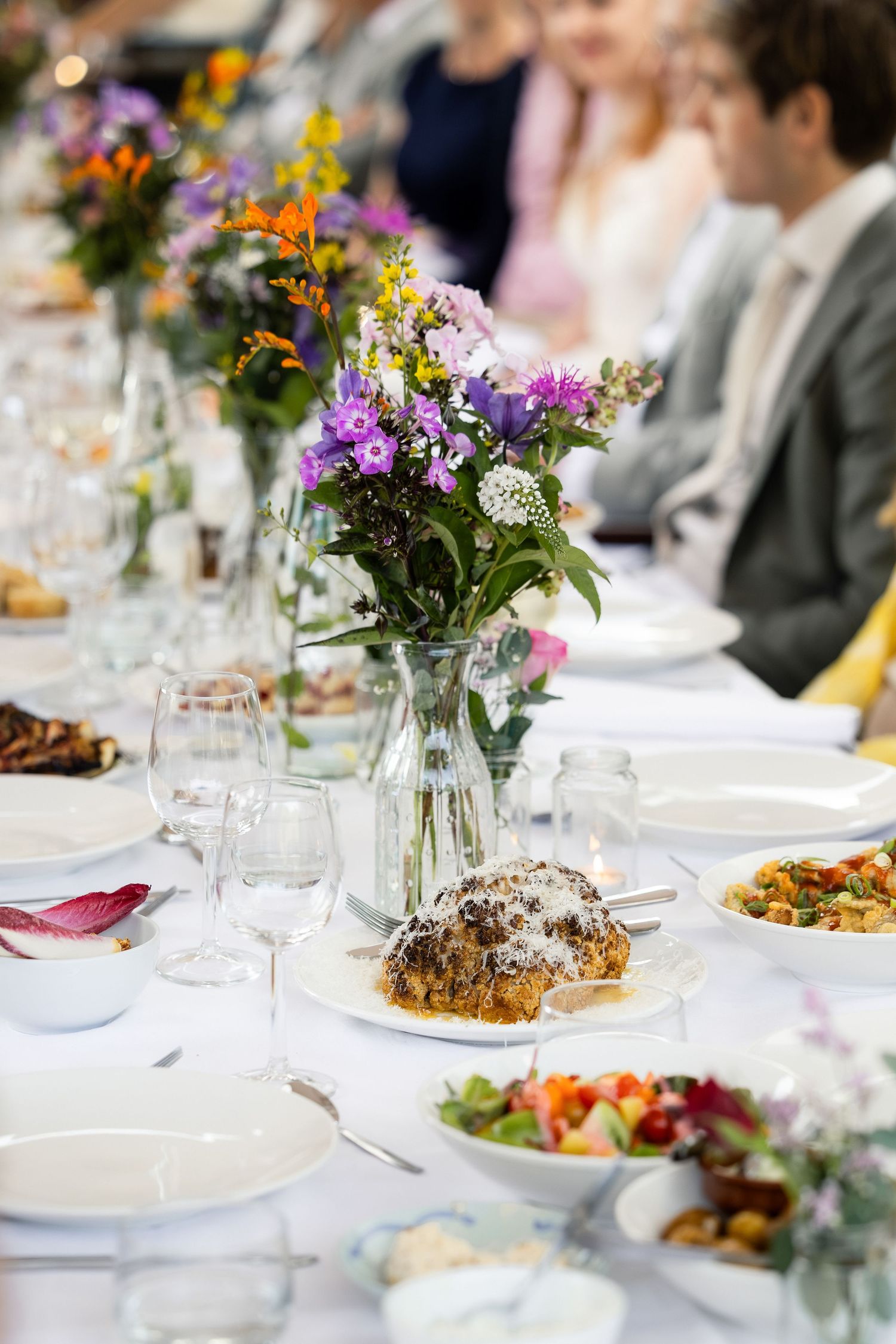 Elegant wedding reception table with wildflower centerpiece, fine glassware, and colorful dishes on white linen.