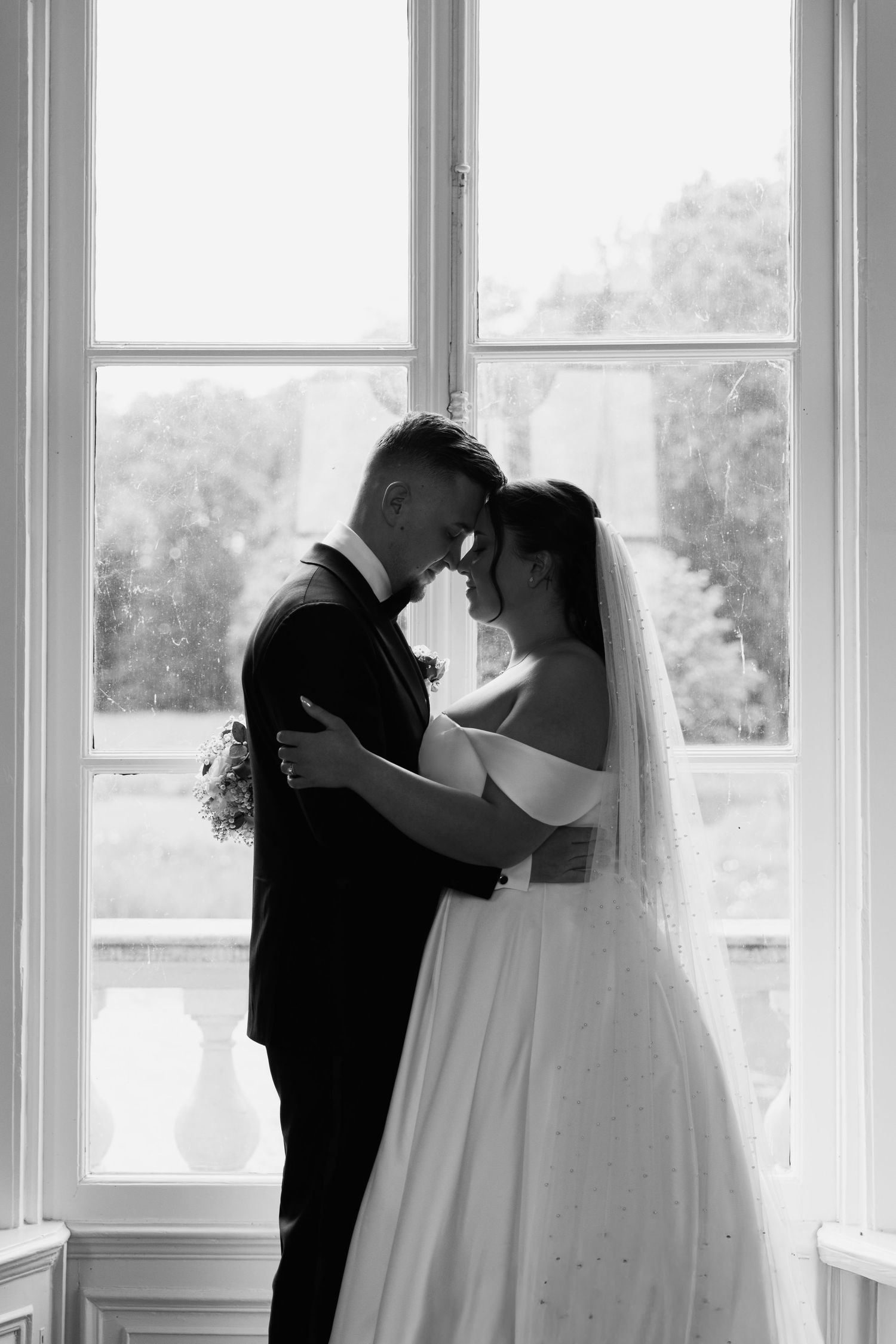 Black and white wedding photo of bride and groom embracing tenderly by a large window.