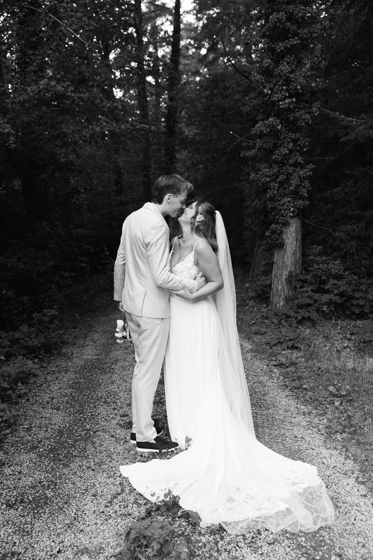 Black and white photo of bride and groom sharing a romantic moment on a forest path, wedding veil trailing behind.