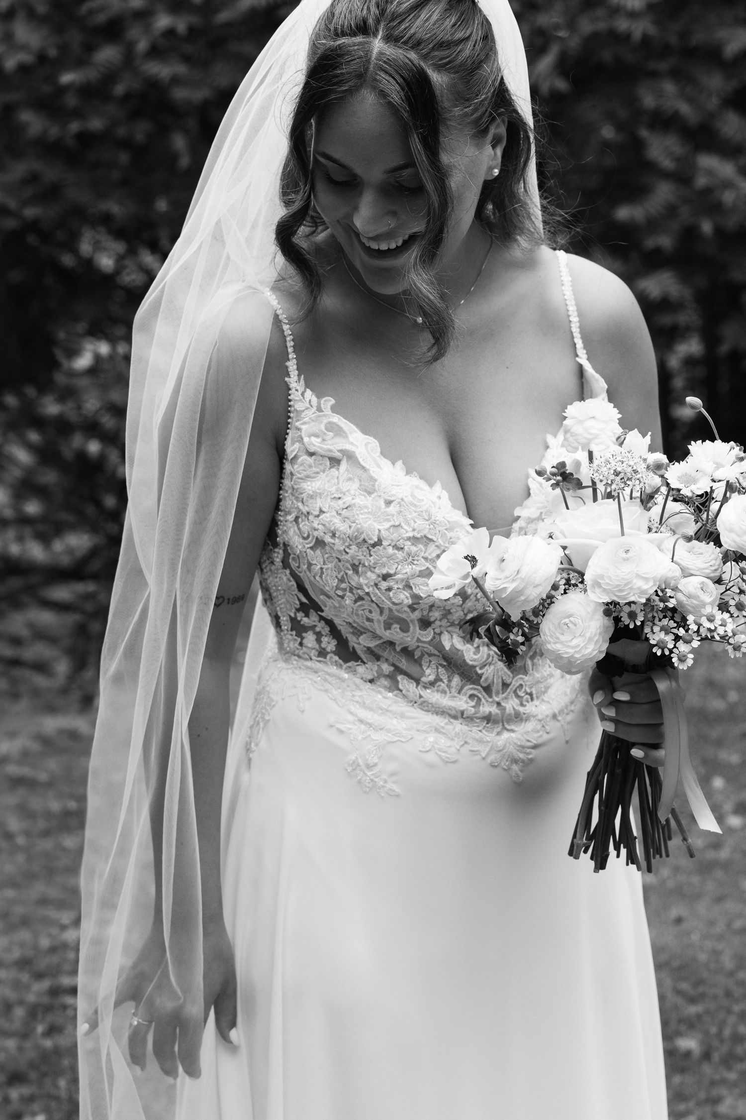 Black and white photo of a laughing bride in lace gown and veil holding a white floral bouquet outdoors.