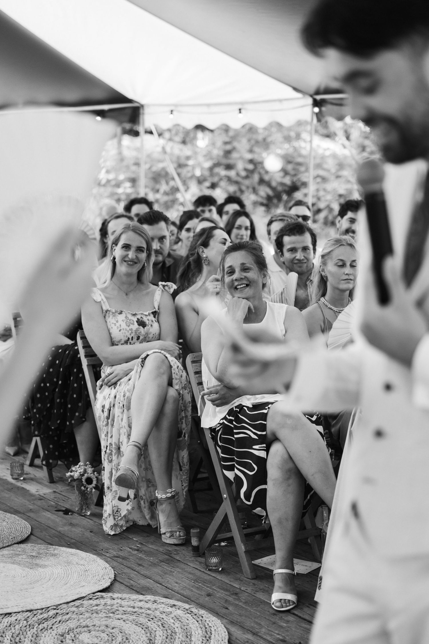 Black and white photo of smiling wedding guests seated outdoors under a tent, listening to a speaker at a summer celebration.