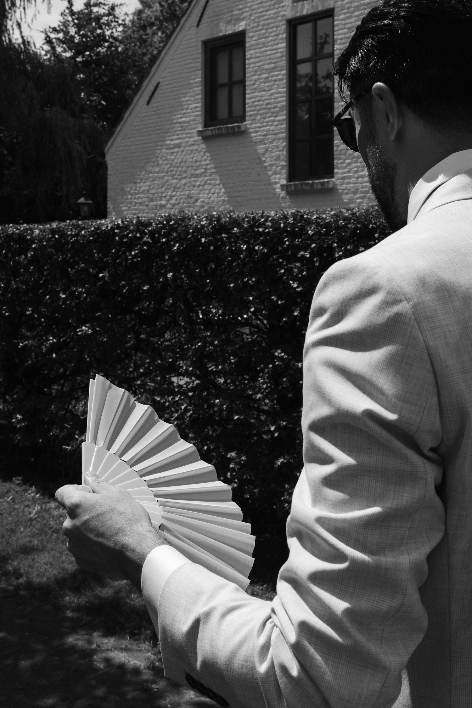 Black and white photo of a man in a light suit holding a decorative hand fan outdoors near a hedge.