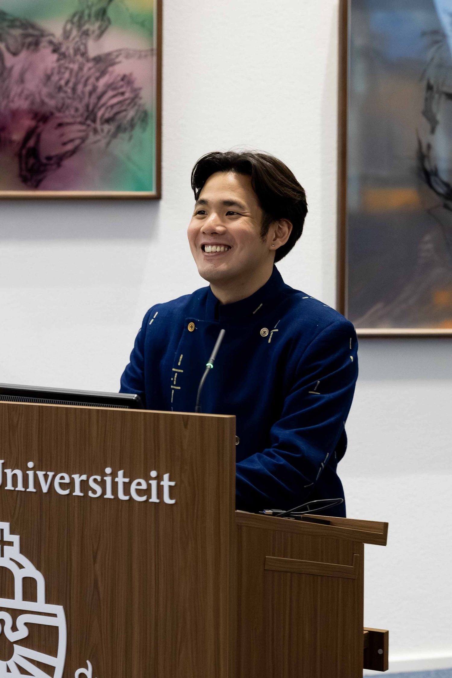 Smiling young man in blue jacket speaking at a university podium labeled Universiteit.
