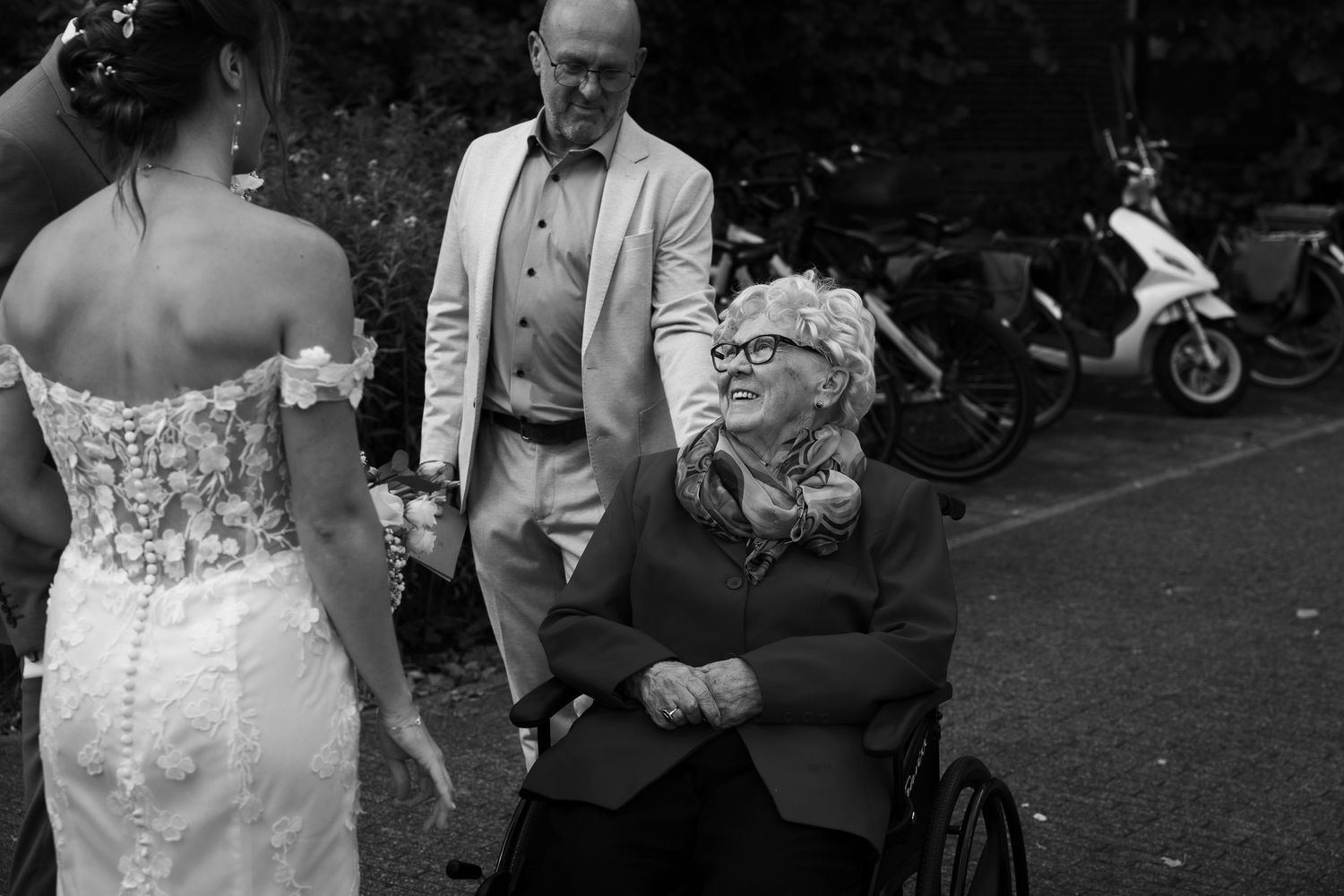 Elderly woman in wheelchair smiling at bride in lace wedding dress at outdoor wedding.