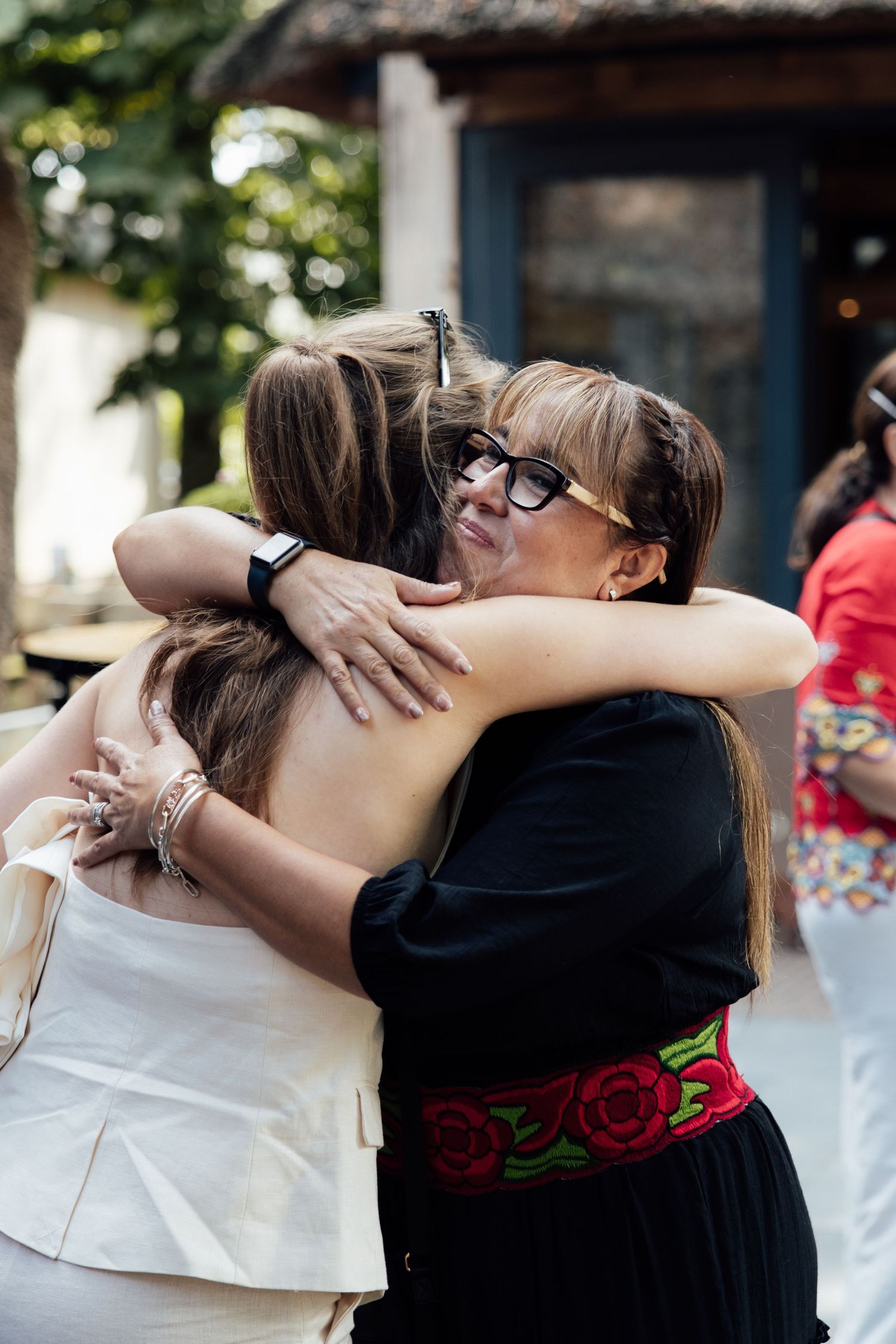 Three women sharing a warm group hug outdoors, one wearing a black dress with red floral embroidered belt.