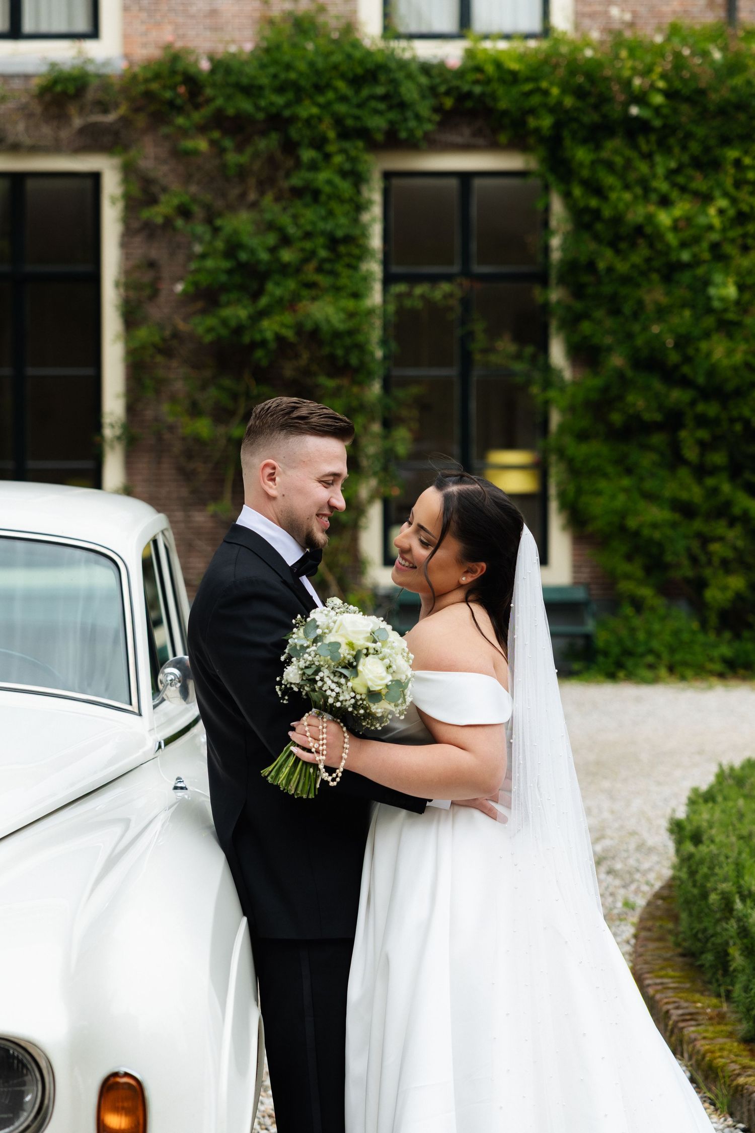 Bride and groom embracing beside a vintage white car in front of an ivy-covered building on their wedding day.