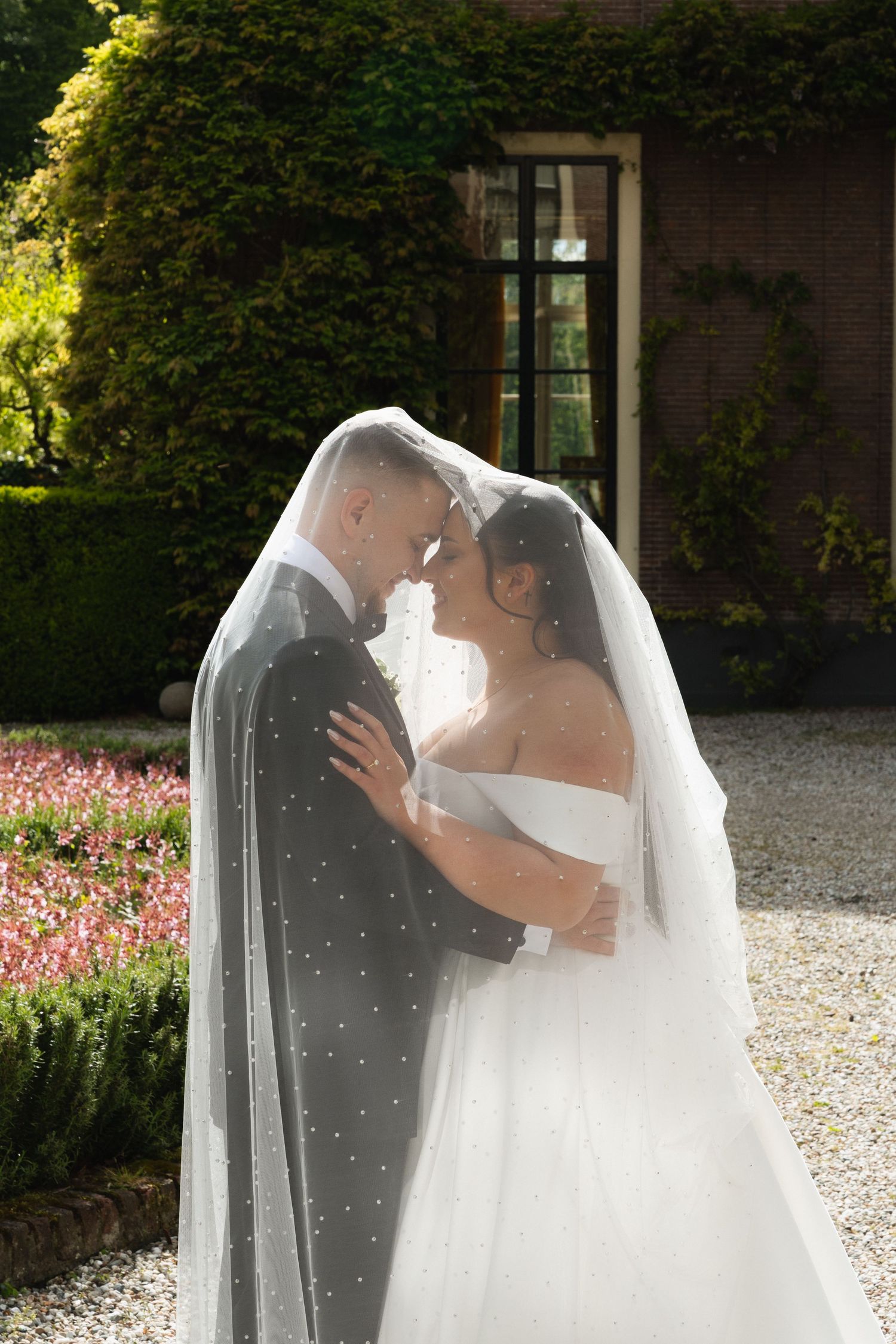 Bride and groom sharing romantic moment under white veil in sunlit garden with brick building backdrop.