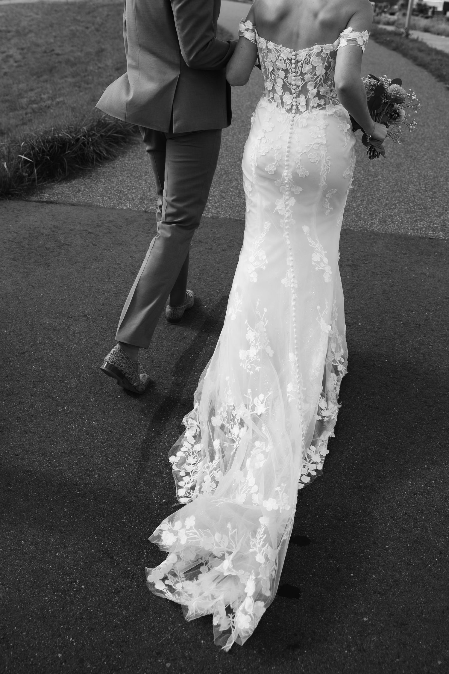 Bride in lace off-shoulder wedding gown with floral train walks with groom in gray suit, black and white photo.