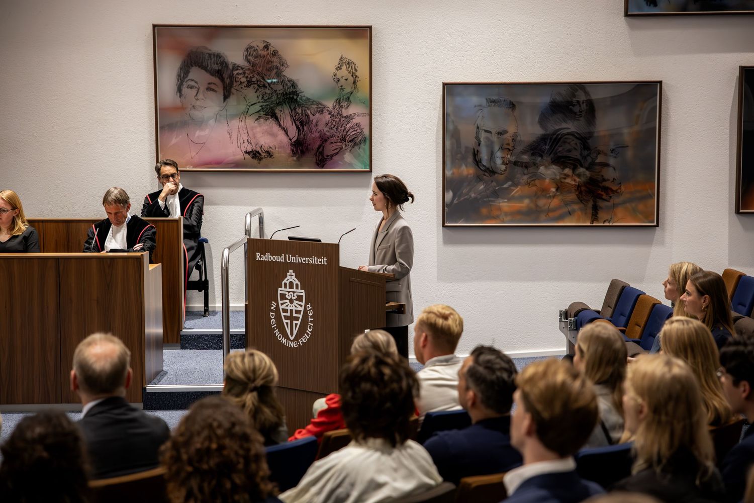 Speaker at Radboud University podium addresses seated audience in formal hall with colorful artwork on walls.