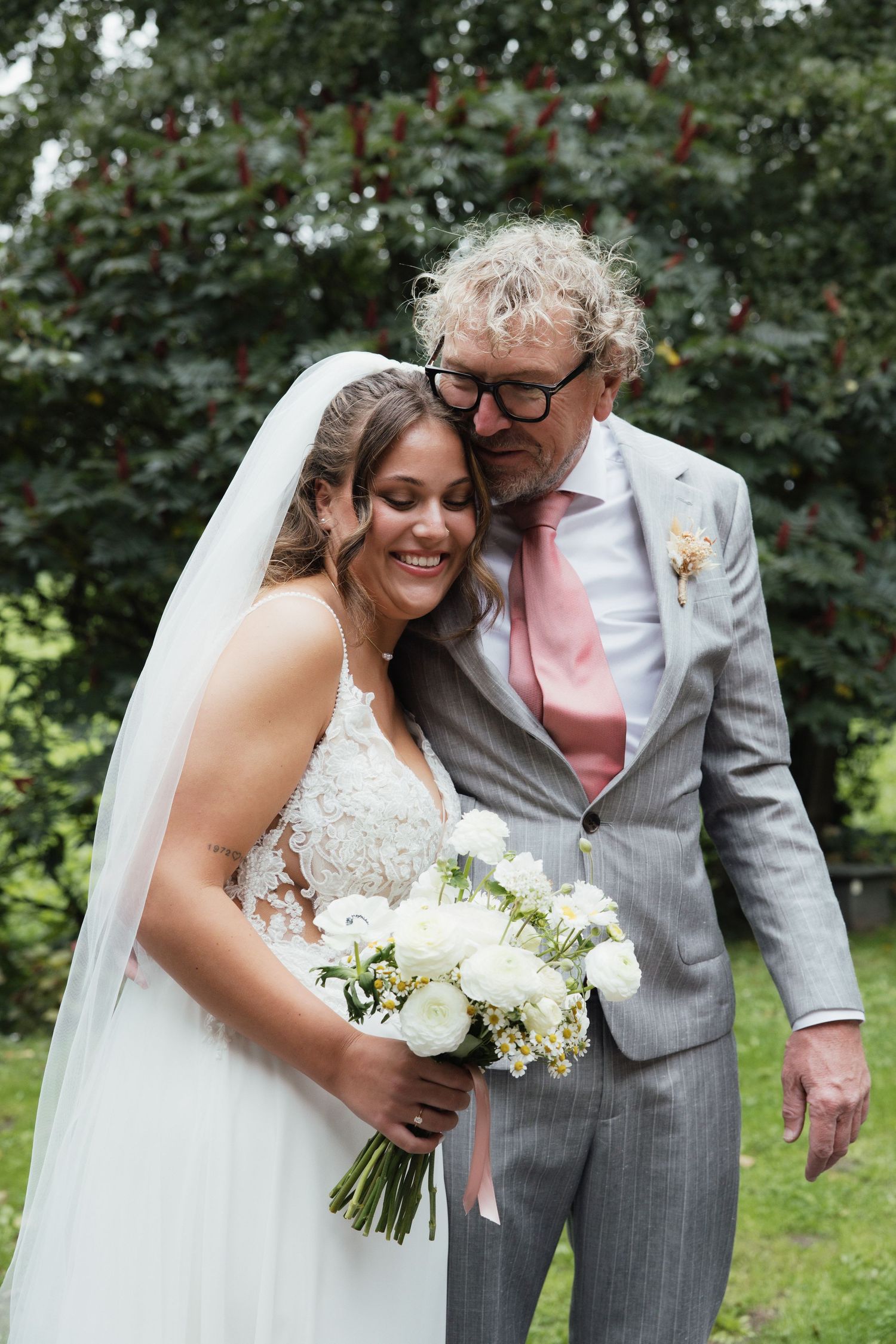 Bride in lace gown holding white bouquet smiles with older man in gray suit and pink tie at outdoor wedding.