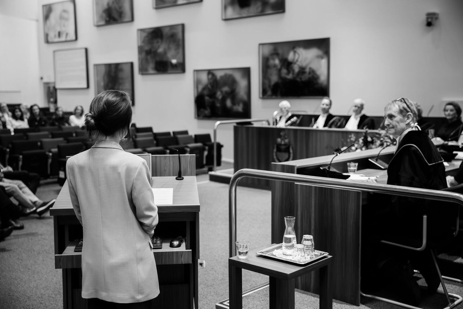Black and white photo of a woman presenting at a podium in a formal courtroom or council chamber setting.