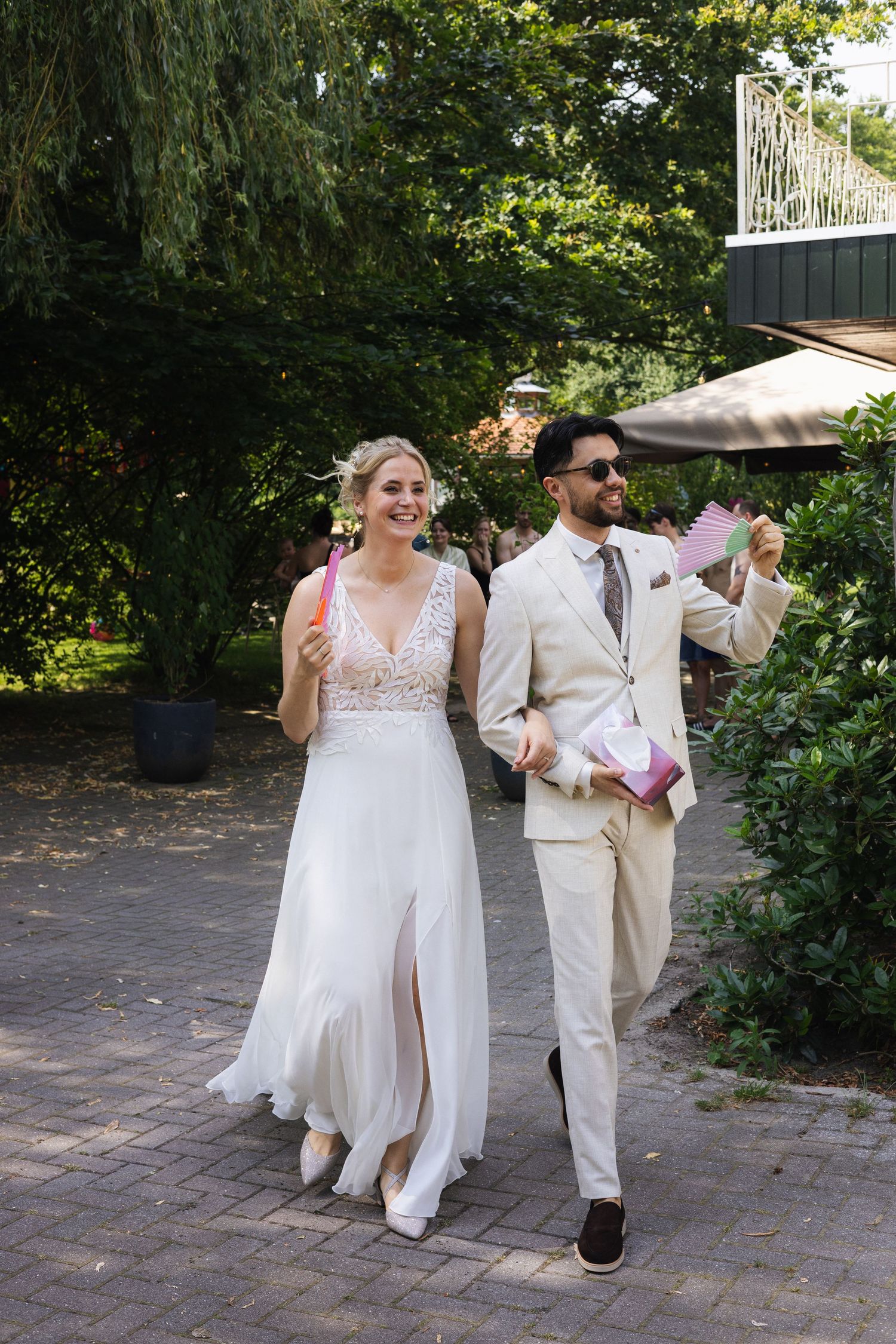 A smiling bride in a white gown and groom in a beige suit walk together outdoors at a sunny wedding venue.