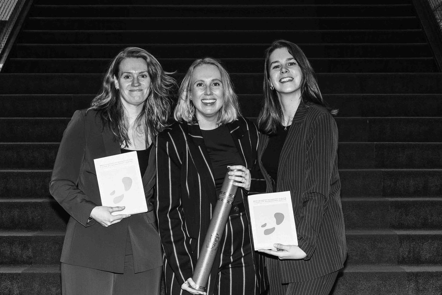 Three women posing on stairs holding books and a trophy in a black and white photo.