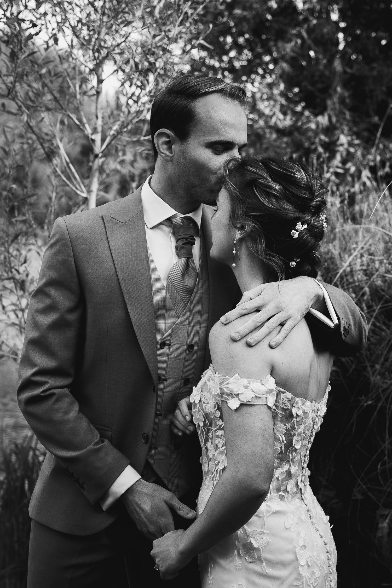 Black and white photo of a groom kissing his bride on the forehead in a garden setting.