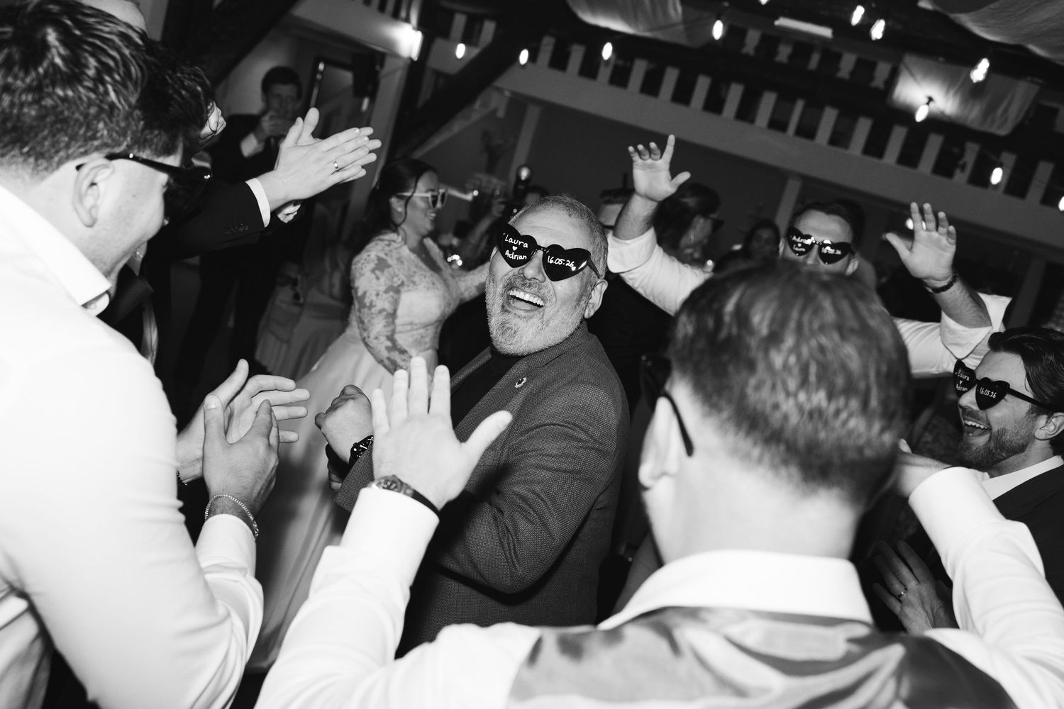Black and white photo of a man in sunglasses smiling at a lively wedding reception dance floor.