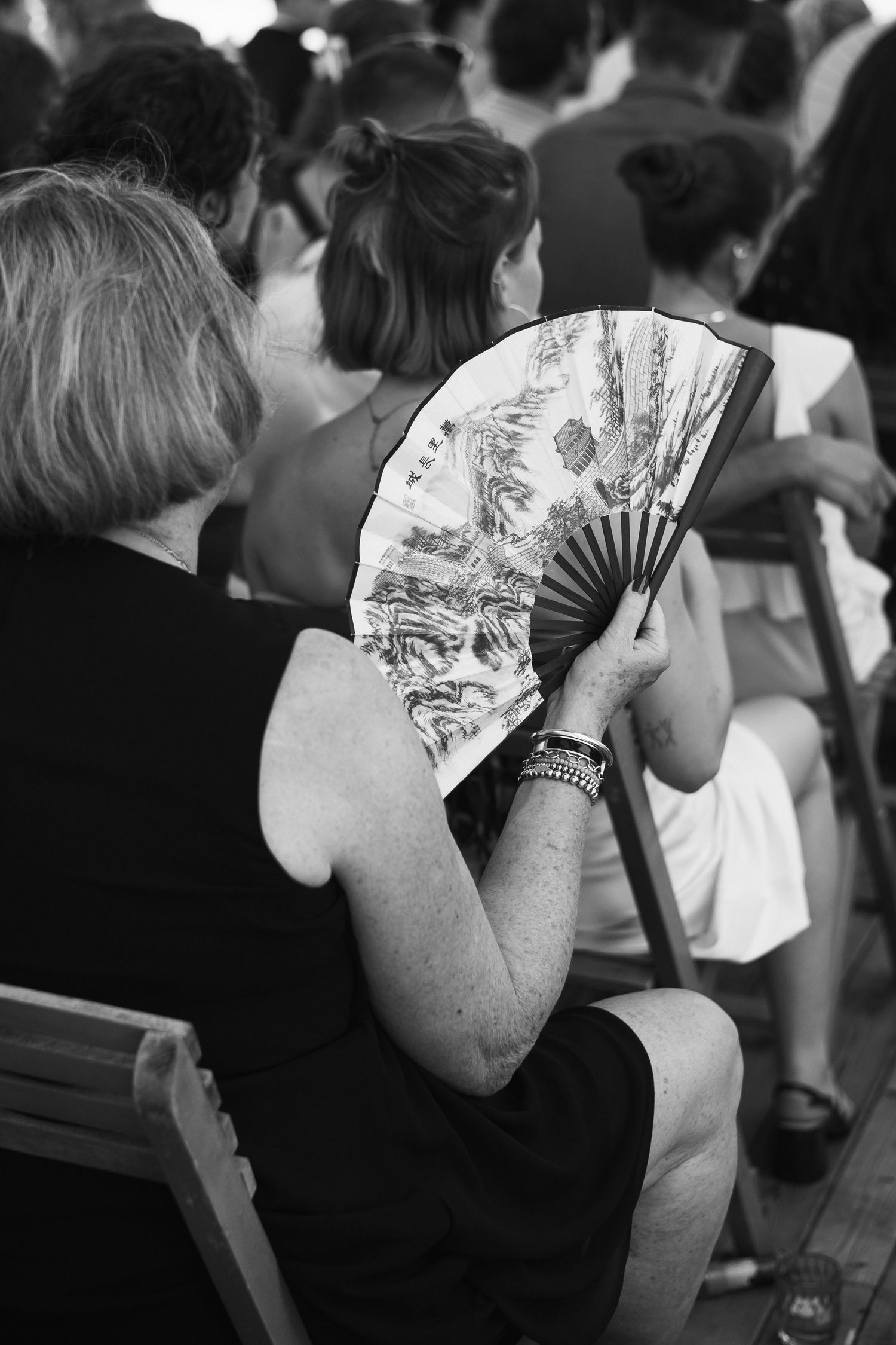 Black and white photo of a woman holding a decorative Asian hand fan in a crowded outdoor seating area.