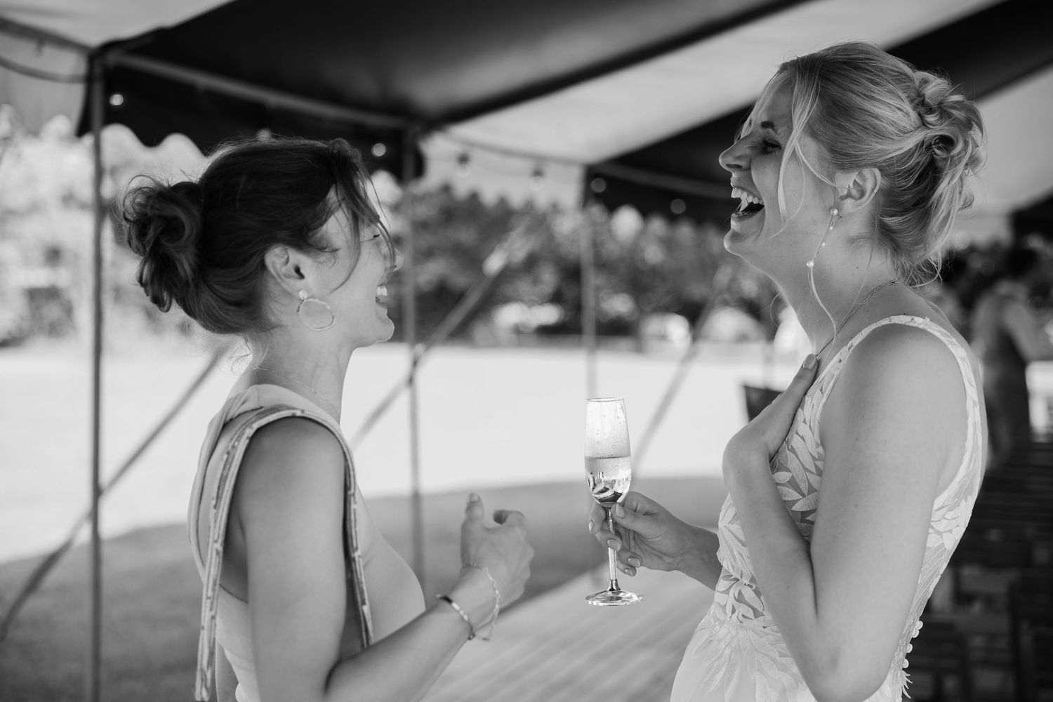 Black and white photo of two women laughing and sharing a joyful moment at a wedding reception under a tent.