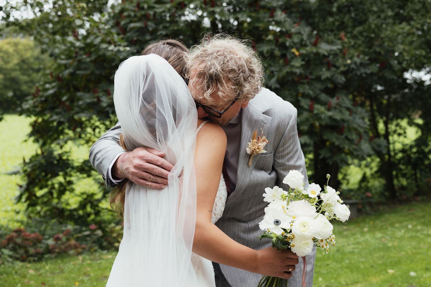 Bride and groom embracing outdoors, bride holding white floral bouquet, wearing veil, groom in gray suit with boutonniere.