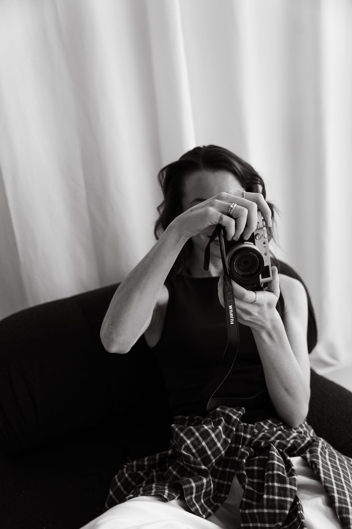 Black and white photo of a woman holding a camera up to her face, sitting on a sofa with a plaid shirt.
