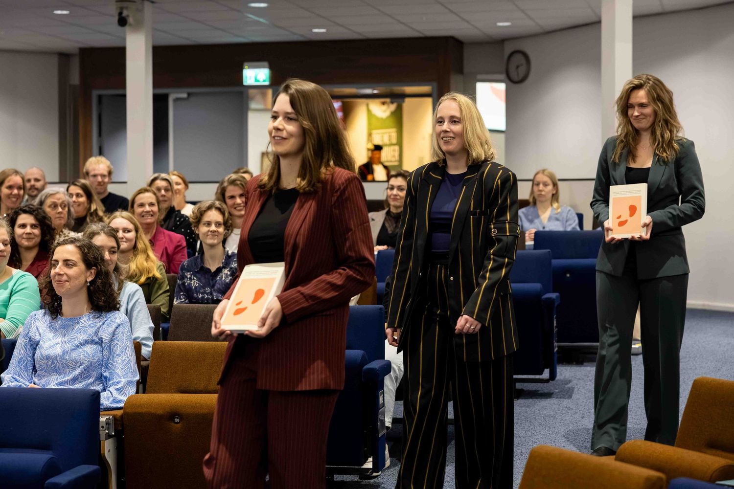 Three women in professional attire walk through a conference room holding books, with an audience seated in rows behind them.