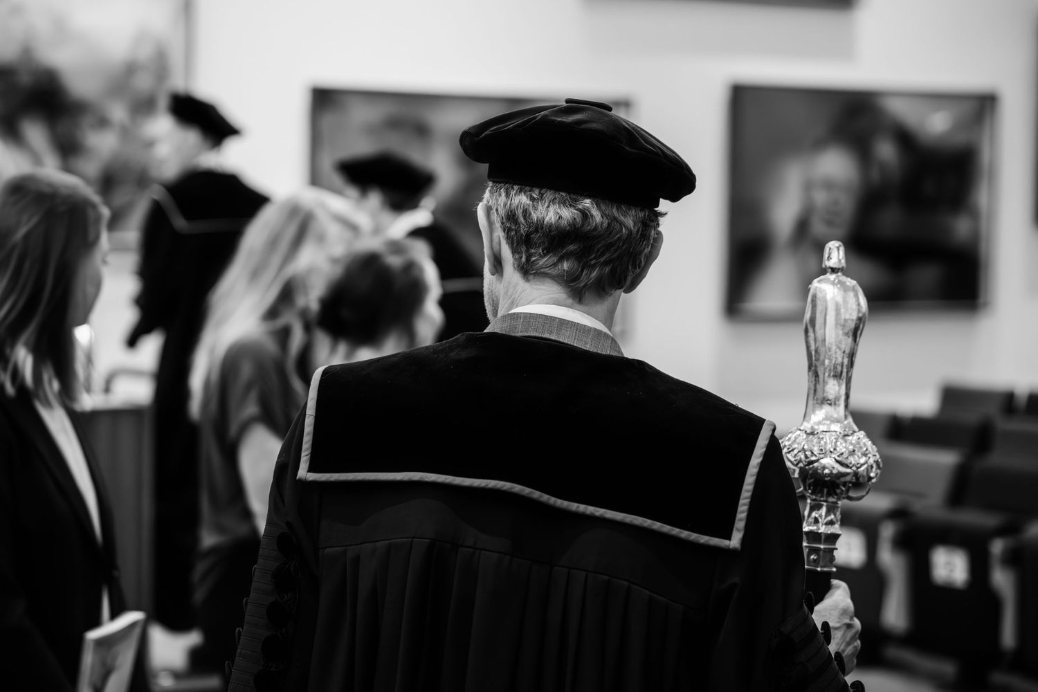 Black and white photo of academic procession with robed figure holding ceremonial mace at formal university graduation ceremony.