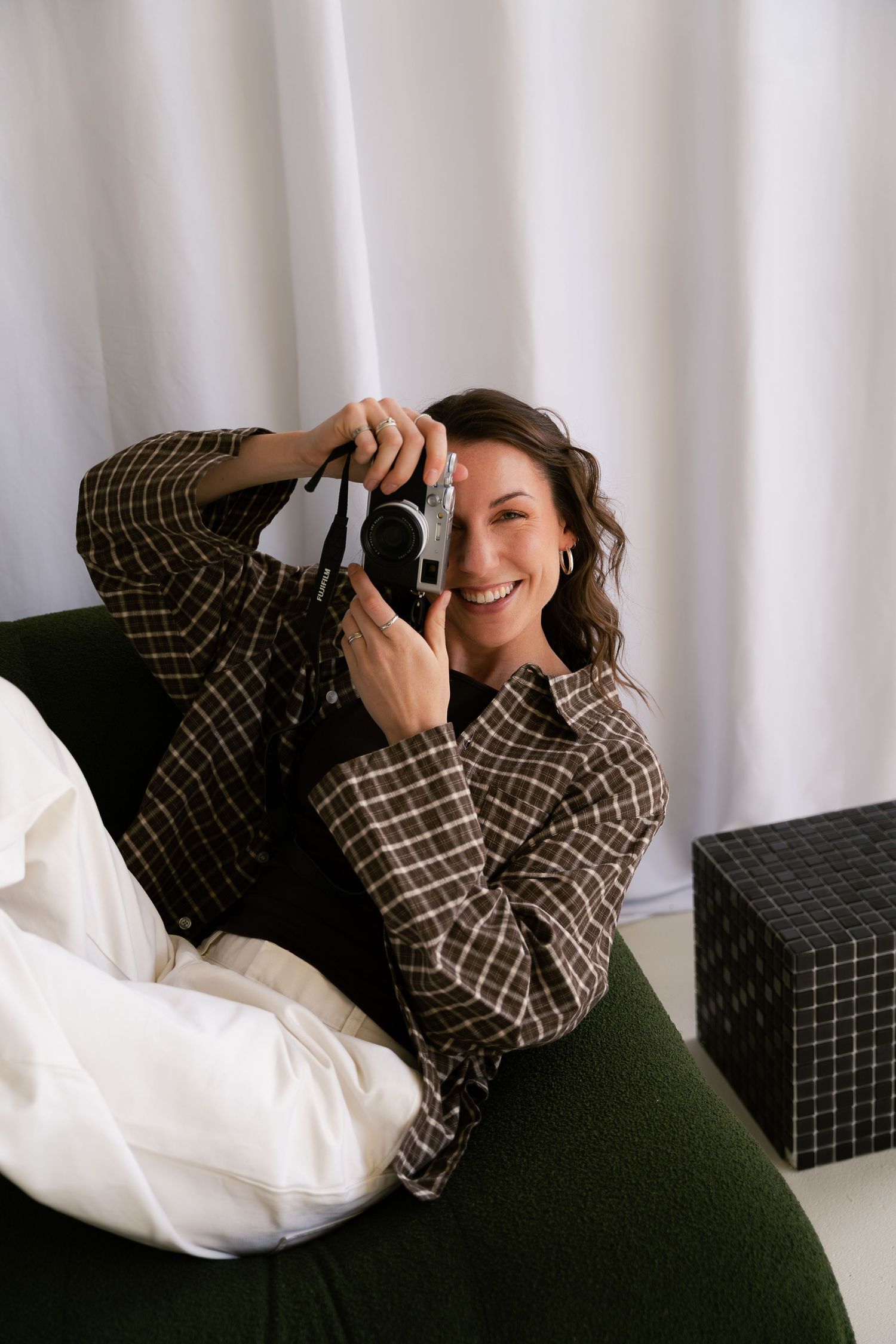 Smiling woman in plaid shirt lying on green sofa holding a vintage film camera indoors.