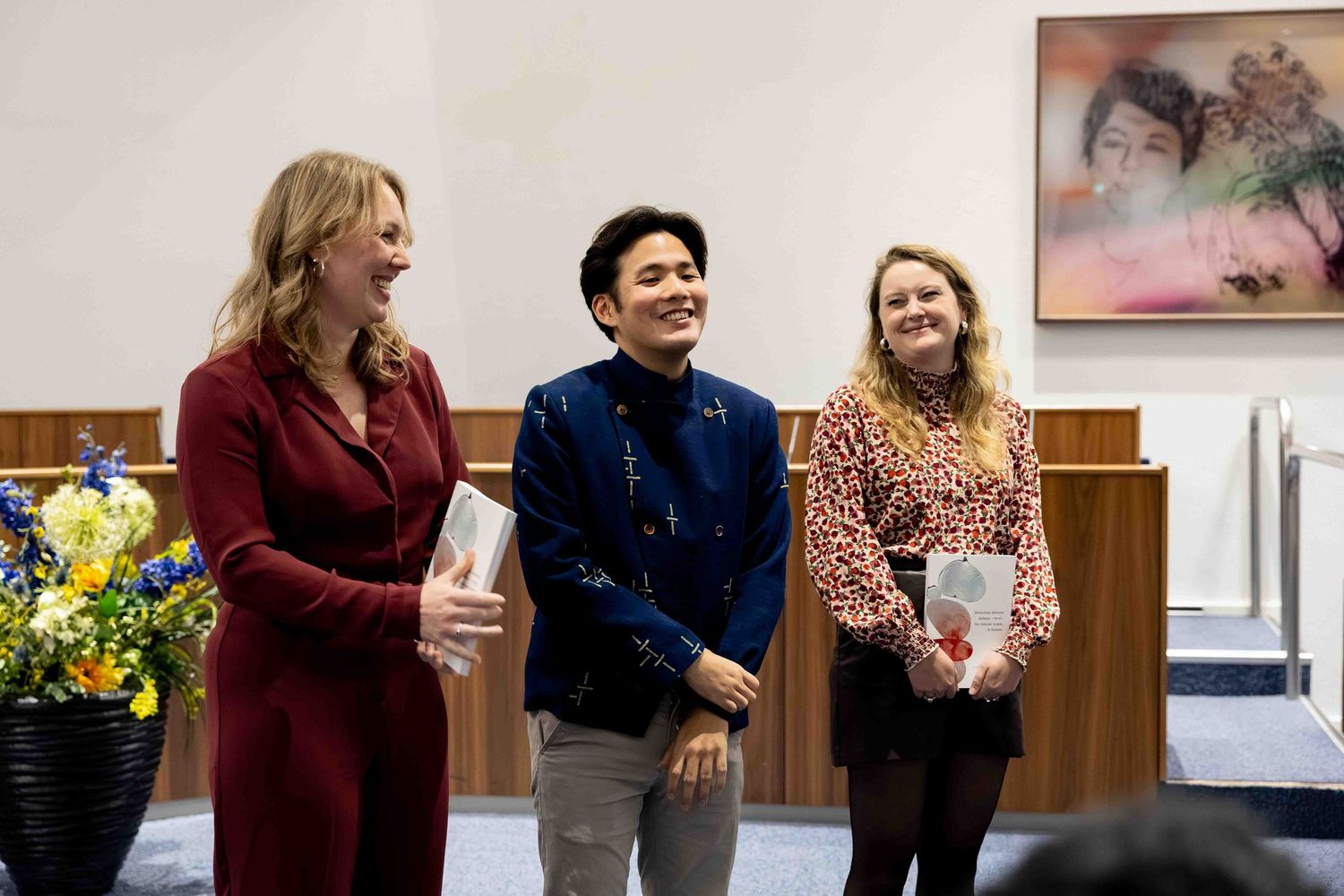 Three people smiling and laughing together at an indoor event, with colorful flowers and artwork in the background.