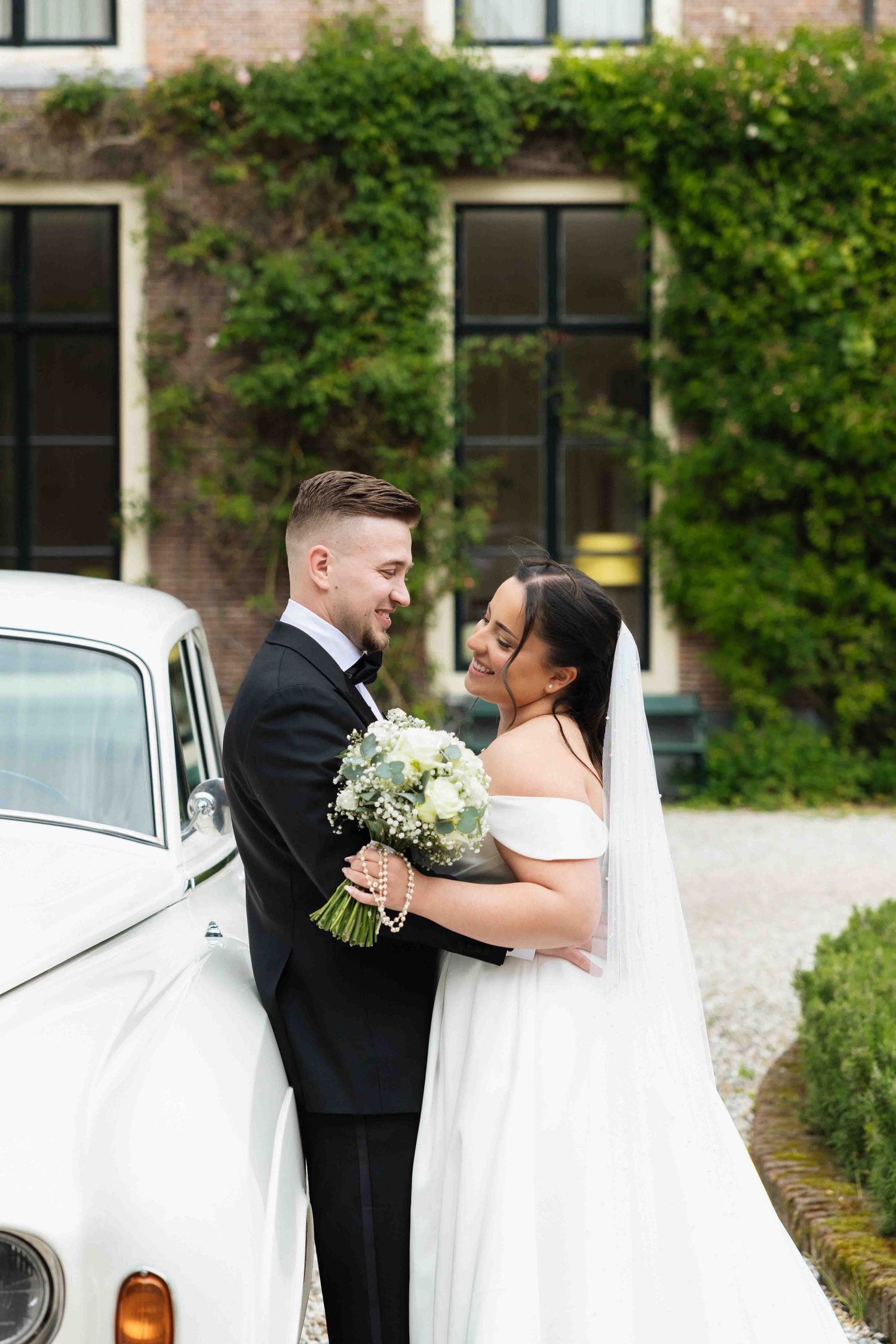Bride and groom embracing near a vintage white car in front of an ivy-covered brick building on their wedding day.