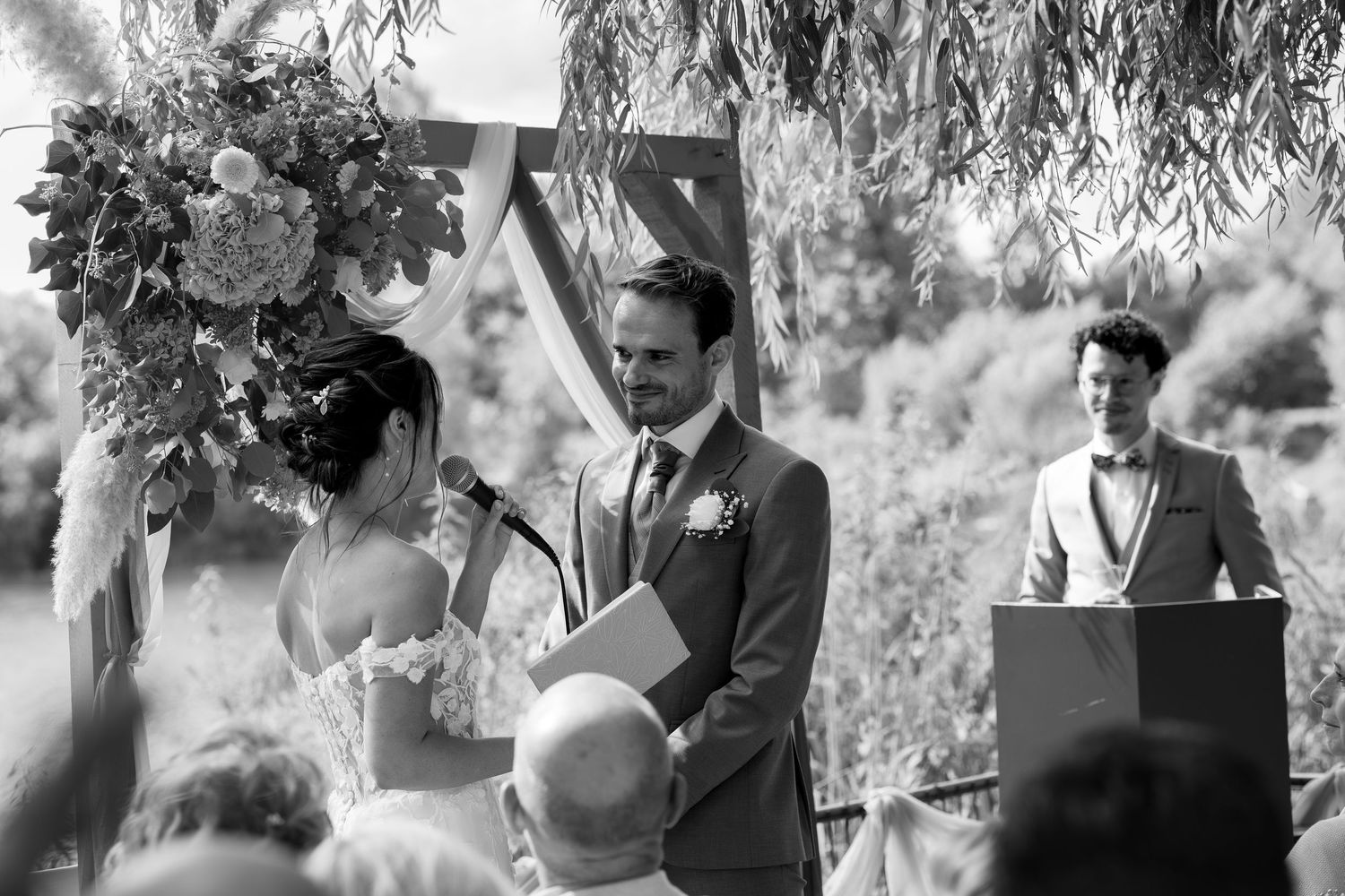 Black and white wedding ceremony photo of bride reading vows to groom outdoors under floral arch with officiant at podium.